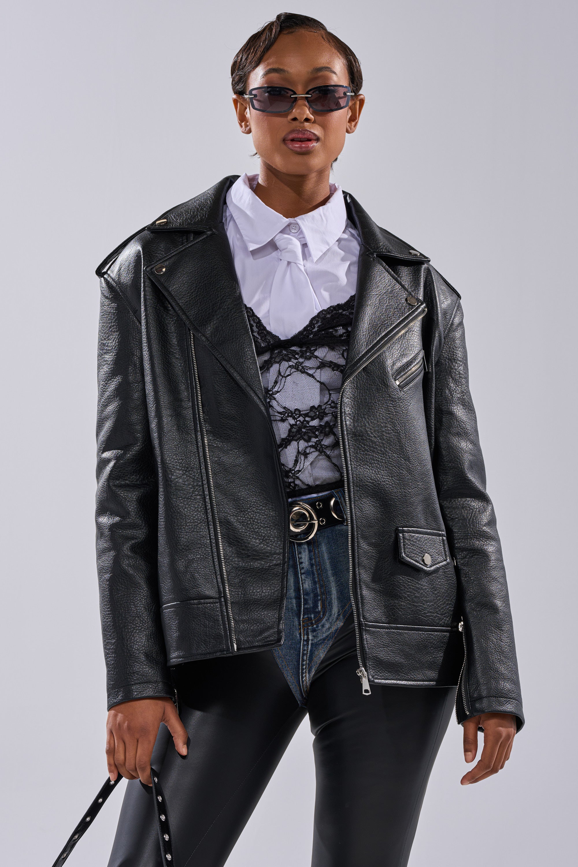 A stylish person wears THE BIGGER THE BETTER OVERSIZED MOTO women's faux leather jacket over a white shirt and black lace corset, paired with sunglasses, blue jeans, and a black belt, holding a strap against a plain light background. 3