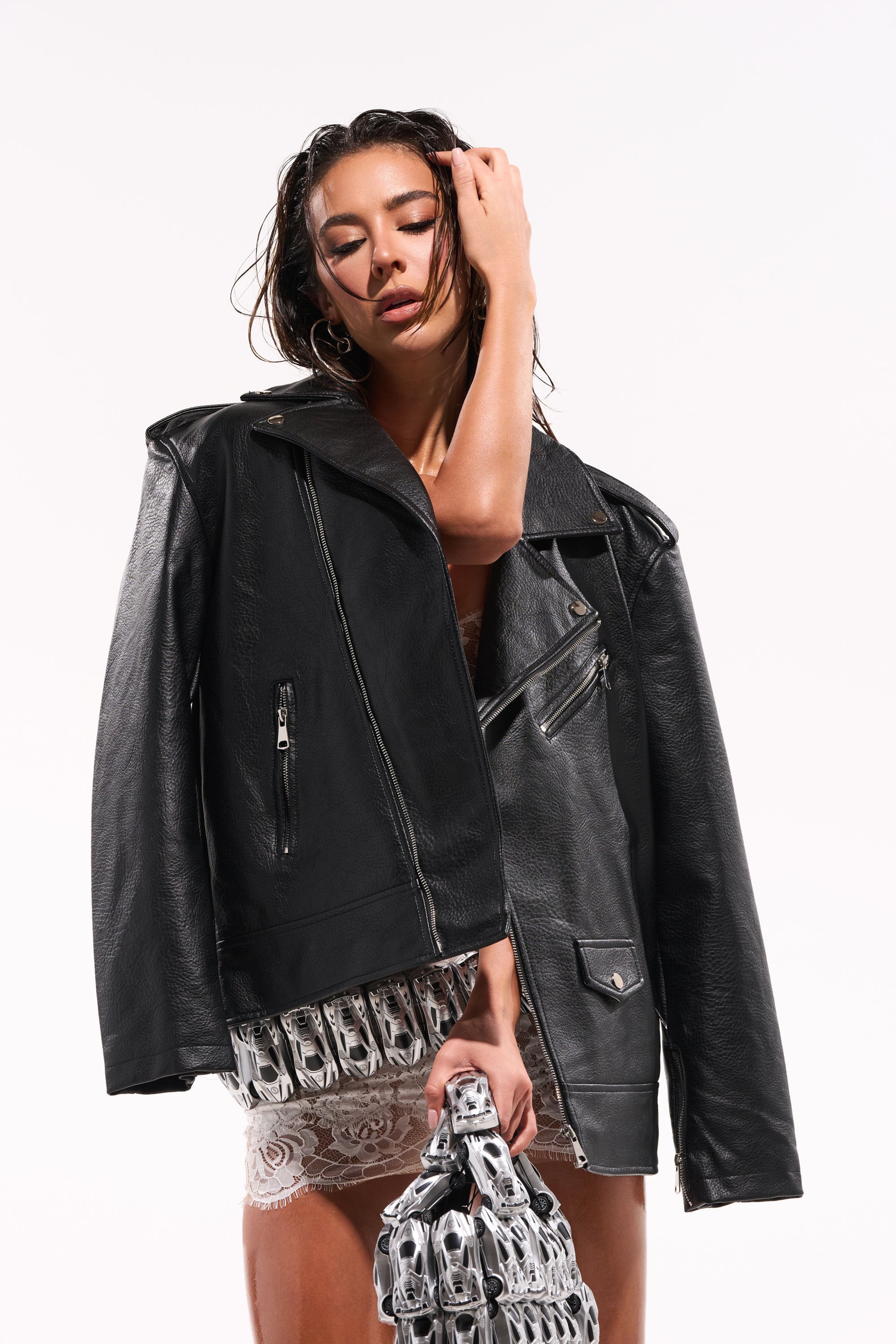 A woman with wet hair poses in THE BIGGER THE BETTER OVERSIZED MOTO jacket over a sheer lace dress, holding a unique silver metallic bag against a plain white background. 0