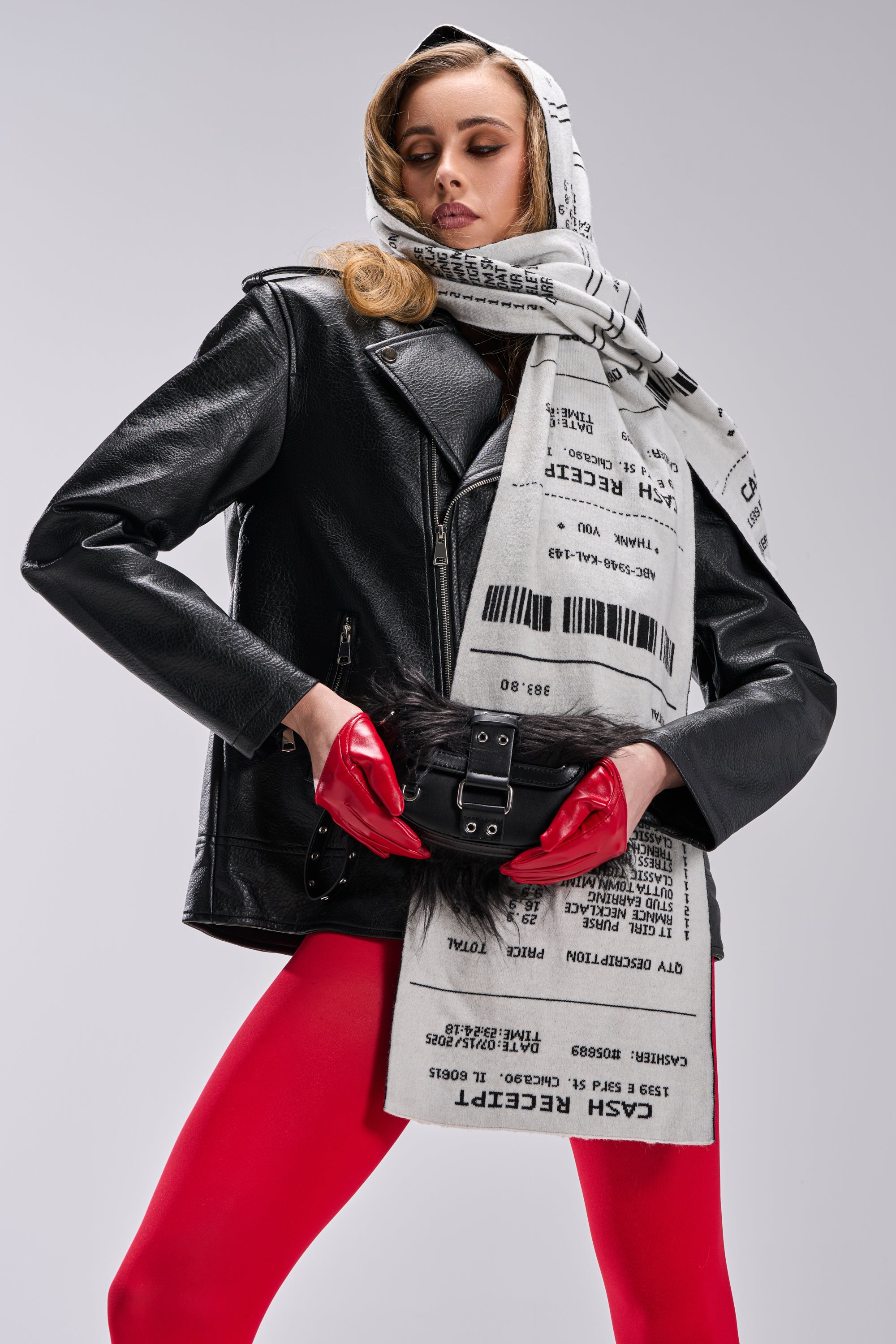 A woman poses in THE BIGGER THE BETTER OVERSIZED MOTO jacket and red tights with gloves. She wears a printed cash receipt scarf on her head and neck, holds a black belt bag, all set against a plain gray background. 5