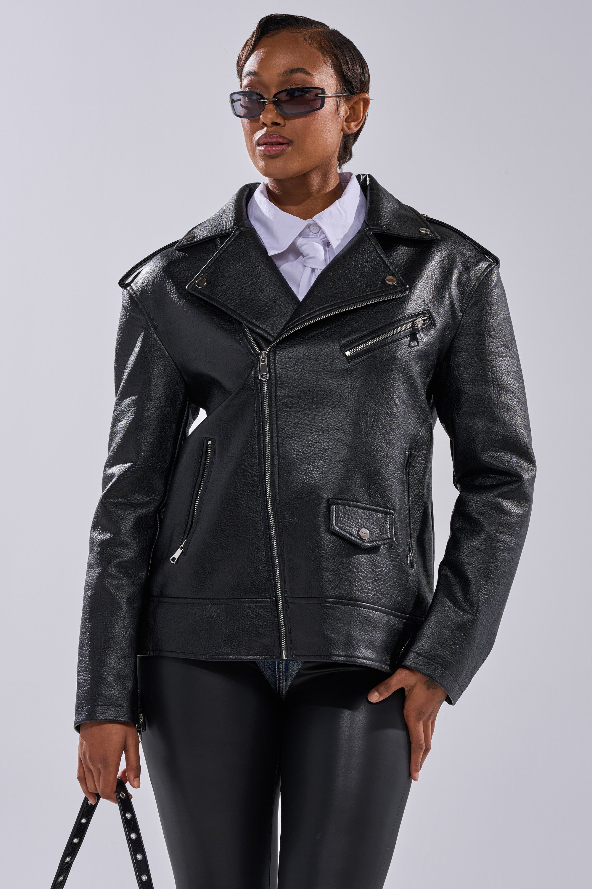 A person in sunglasses, a white collared shirt, black pants, and THE BIGGER THE BETTER OVERSIZED MOTO jacket stands against a plain light gray background, holding a black strap in one hand. 4