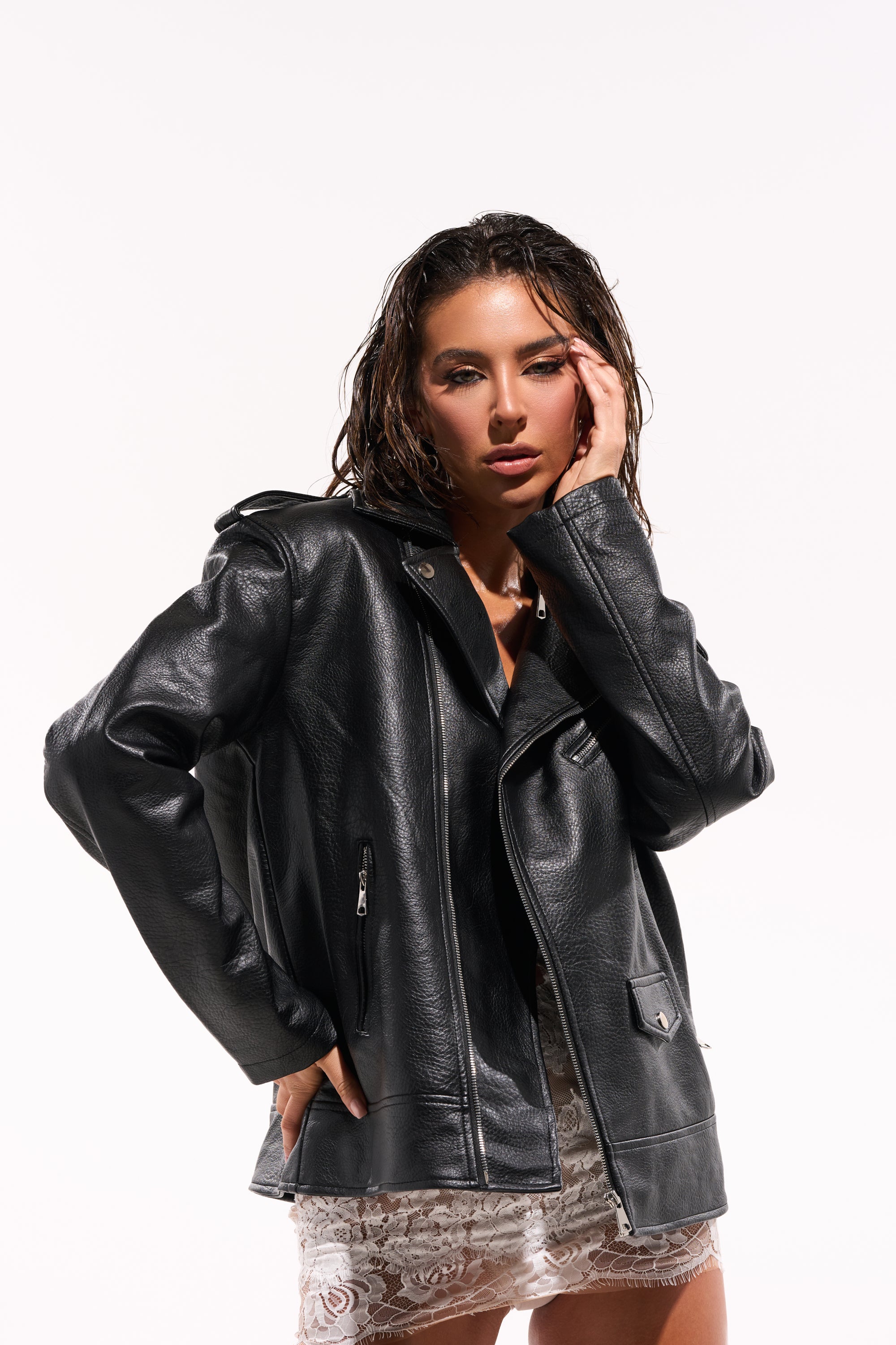 A woman with wet hair poses confidently with one hand on her cheek, wearing THE BIGGER THE BETTER OVERSIZED MOTO over a white lace dress against a plain white background. 1
