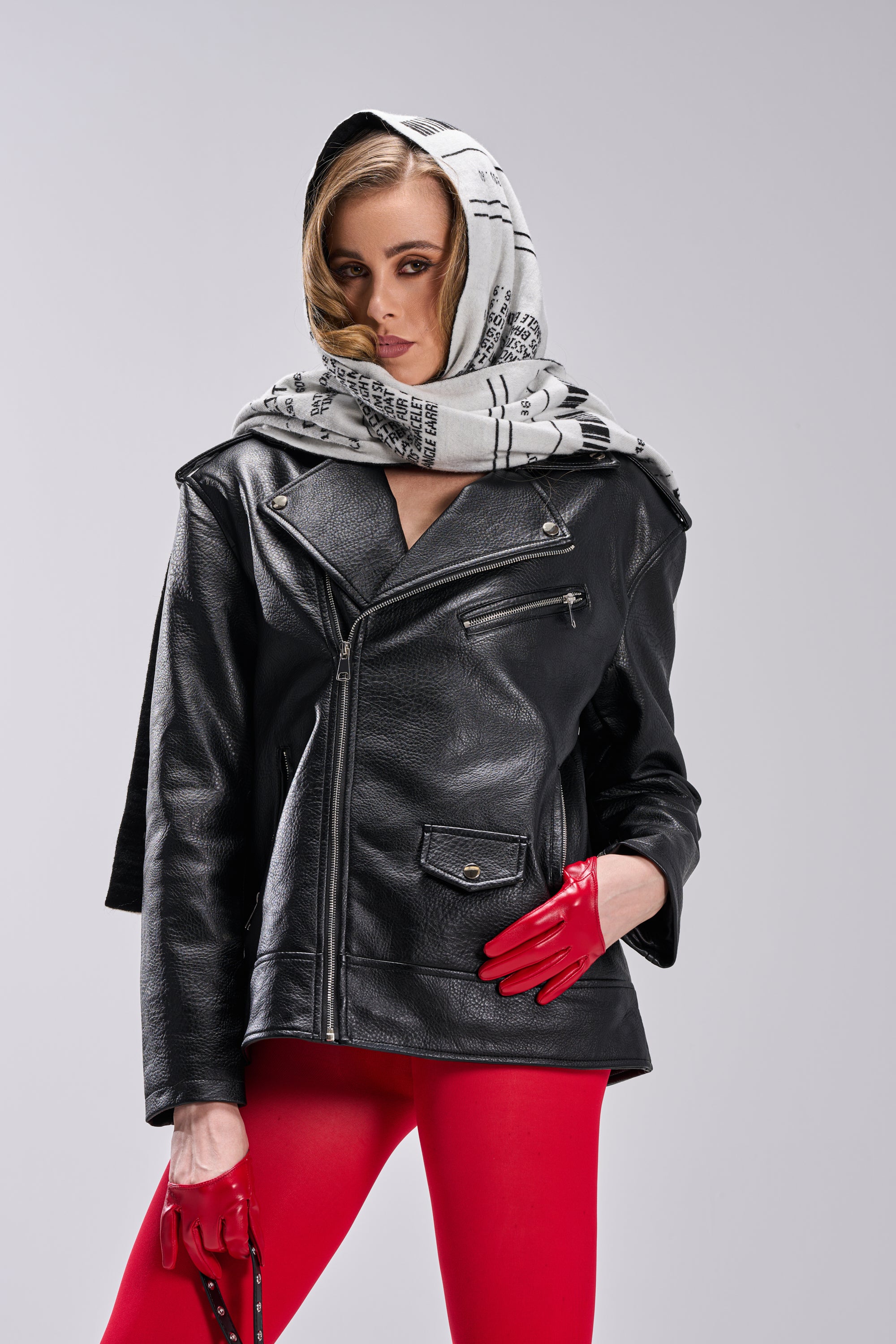 A stylish person poses confidently in THE BIGGER THE BETTER OVERSIZED MOTO jacket, red gloves and tights, and a light scarf with black patterns wrapped around their head. 6
