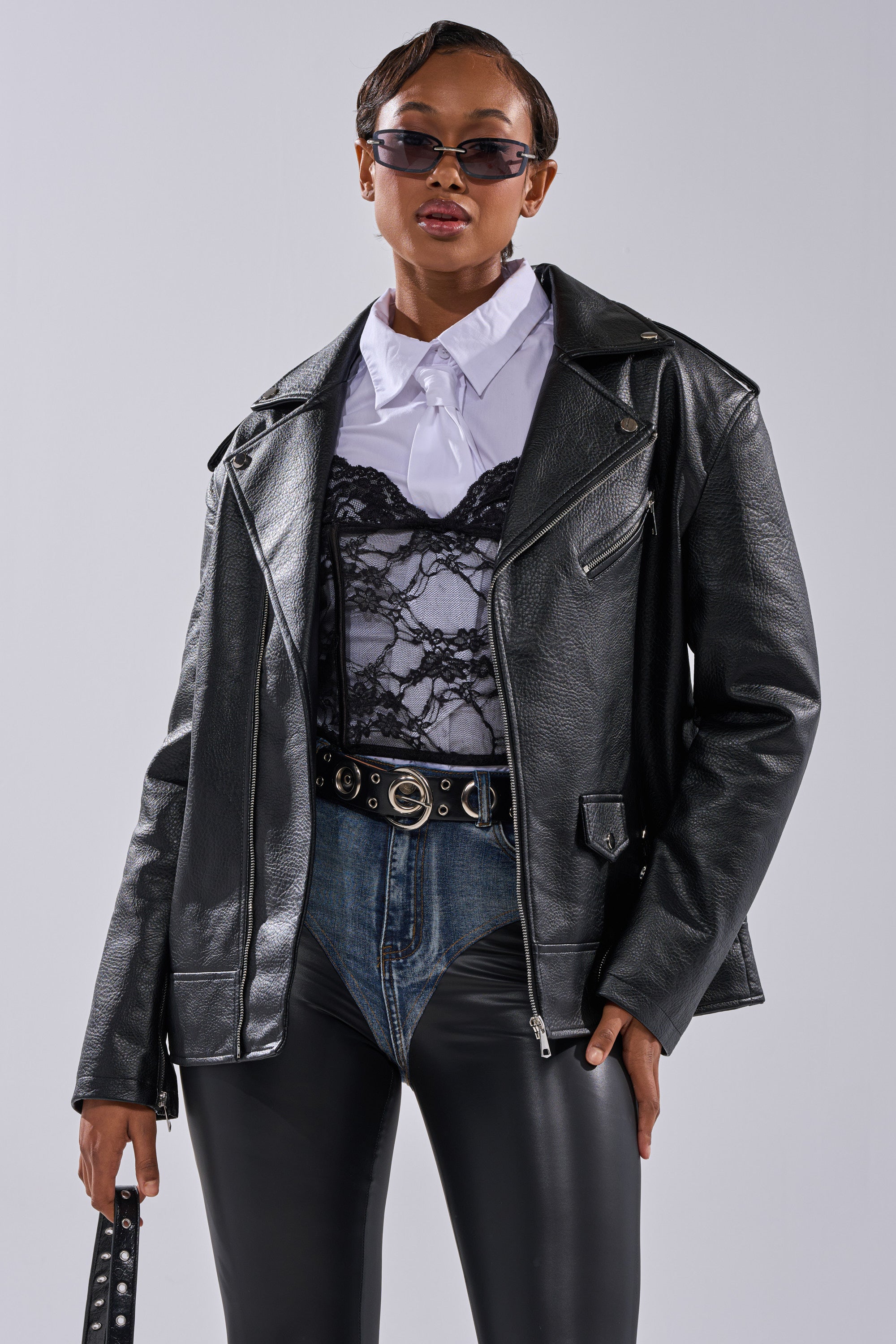 A stylish person wearing THE BIGGER THE BETTER OVERSIZED MOTO jacket over a white collared shirt, lace top, dark sunglasses, high-waisted denim shorts, black belt, and black leather pants stands against a plain background. 10