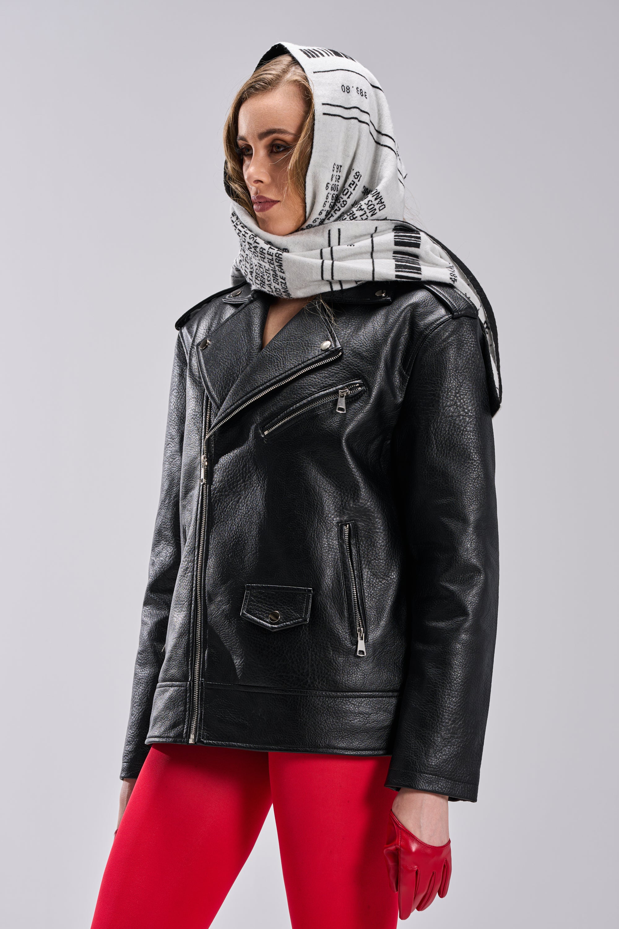 A person wears THE BIGGER THE BETTER OVERSIZED MOTO jacket, red pants, red gloves, and a white scarf with black text and patterns while standing against a plain light gray background. 7