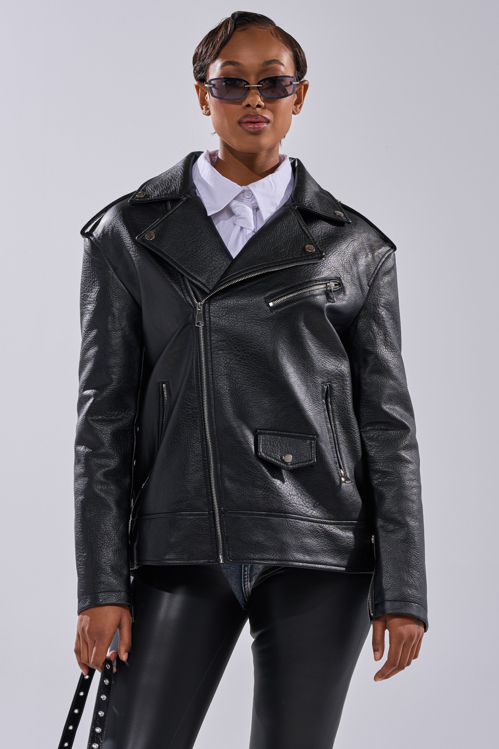 A person in sunglasses, a white collared shirt, black pants, and THE BIGGER THE BETTER OVERSIZED MOTO stands against a plain light background, holding a black strap with silver studs. 11