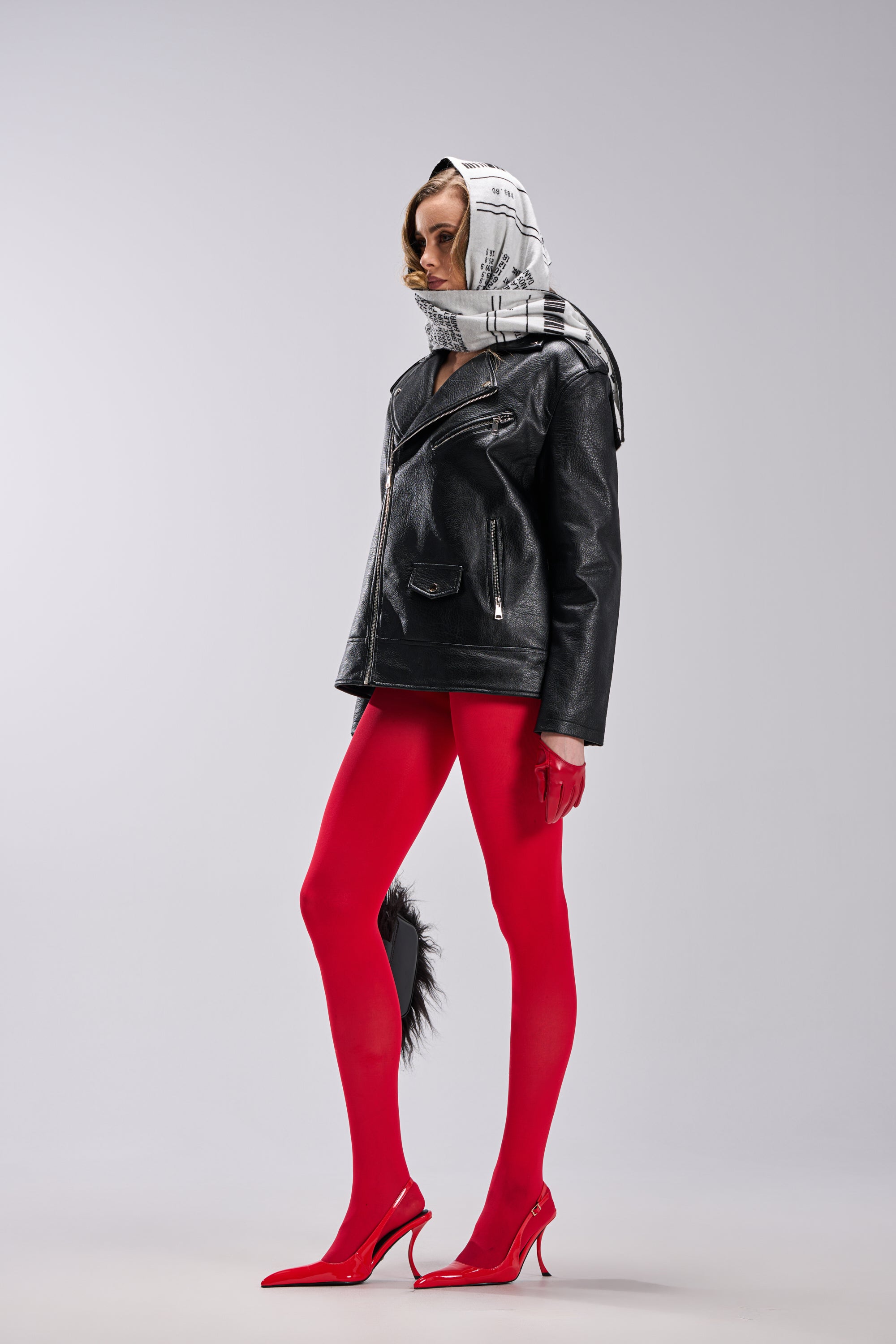 A person stands in profile wearing THE BIGGER THE BETTER OVERSIZED MOTO black faux leather jacket, red tights, red pointed heels, red gloves, and a white patterned headscarf against a plain light gray background. 8