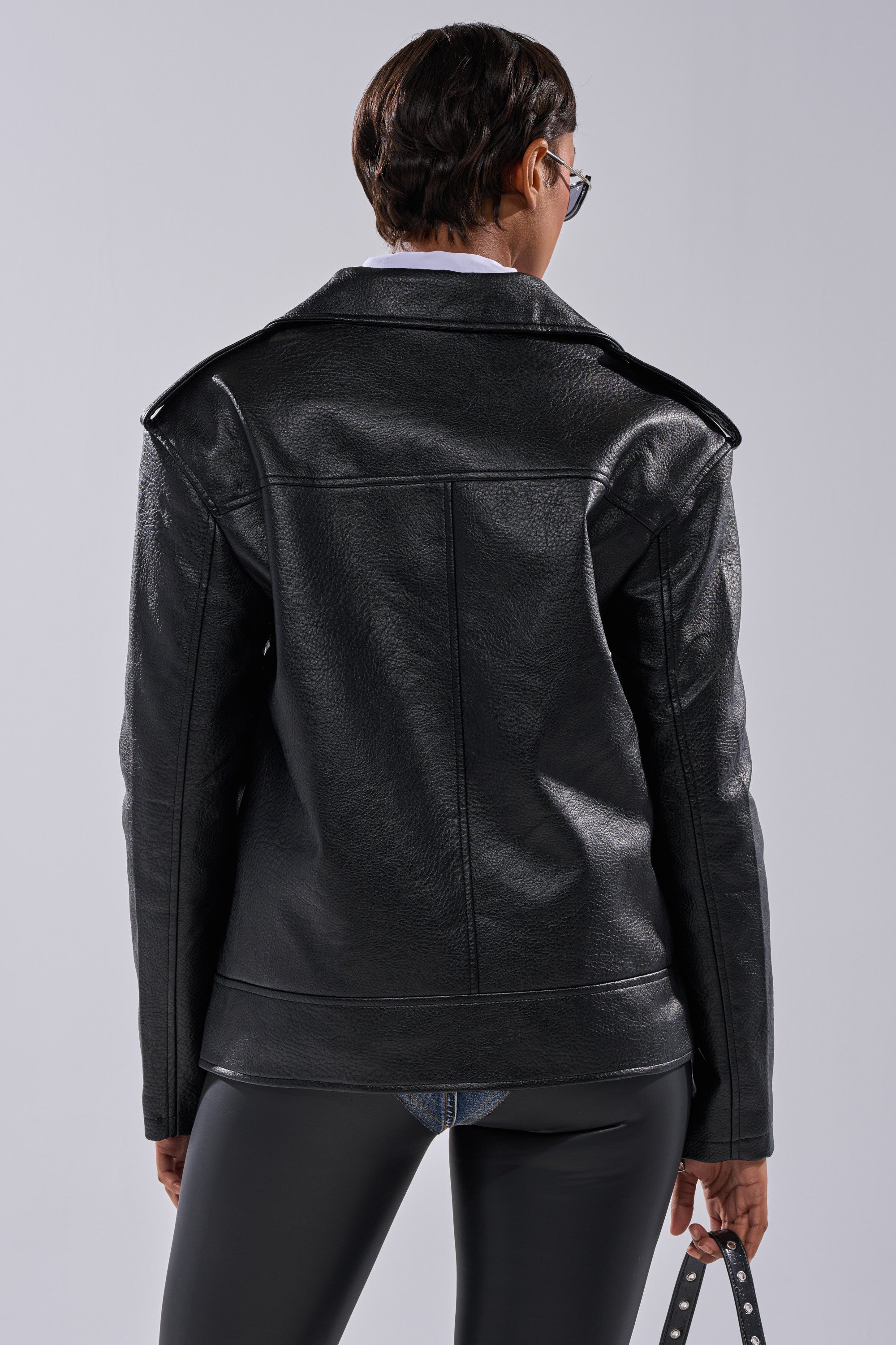 A person with short dark hair and sunglasses is shown from the back wearing THE BIGGER THE BETTER OVERSIZED MOTO jacket and black pants, holding a strap in their right hand against a plain light background. 13
