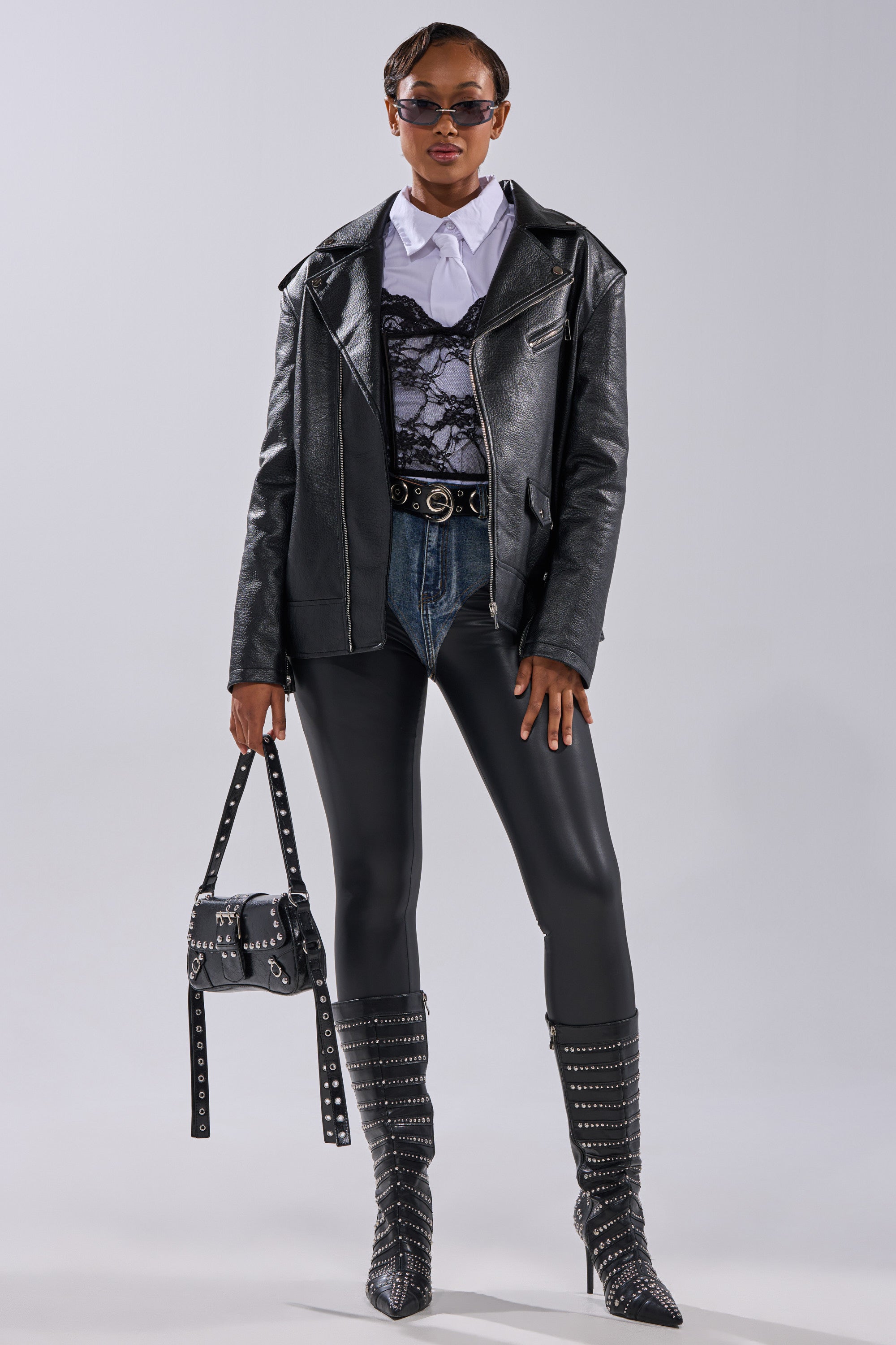 A person stands confidently in THE BIGGER THE BETTER OVERSIZED MOTO jacket, paired with a white shirt, lace top, high-waisted jeans, black studded boots, sunglasses, and a matching studded handbag against a plain light gray background. 15