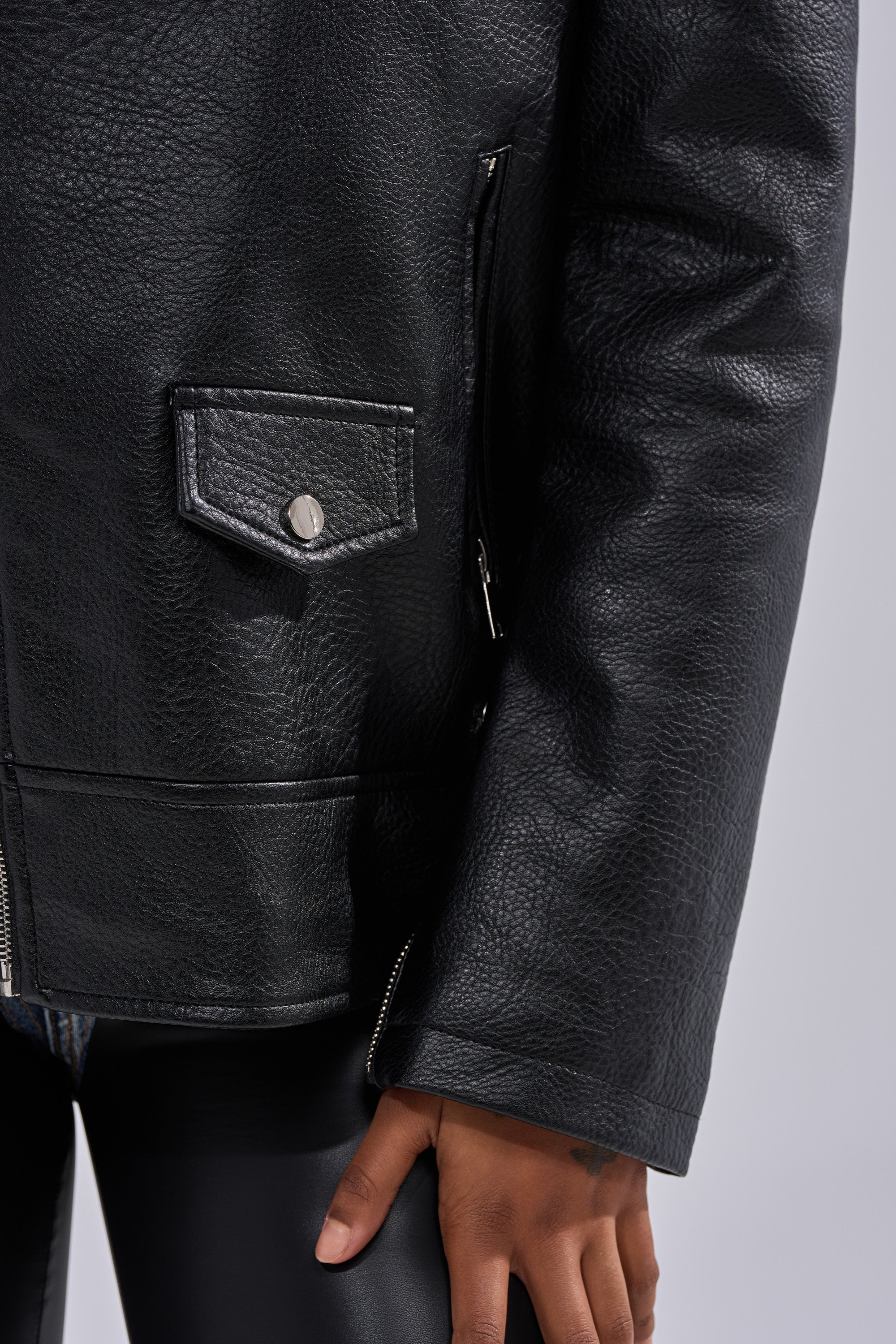 A close-up of a person wearing THE BIGGER THE BETTER OVERSIZED MOTO, a textured black leather jacket with zip and buttoned pocket, paired with shiny black pants. Their left hand appears near the bottom right of the image. 16