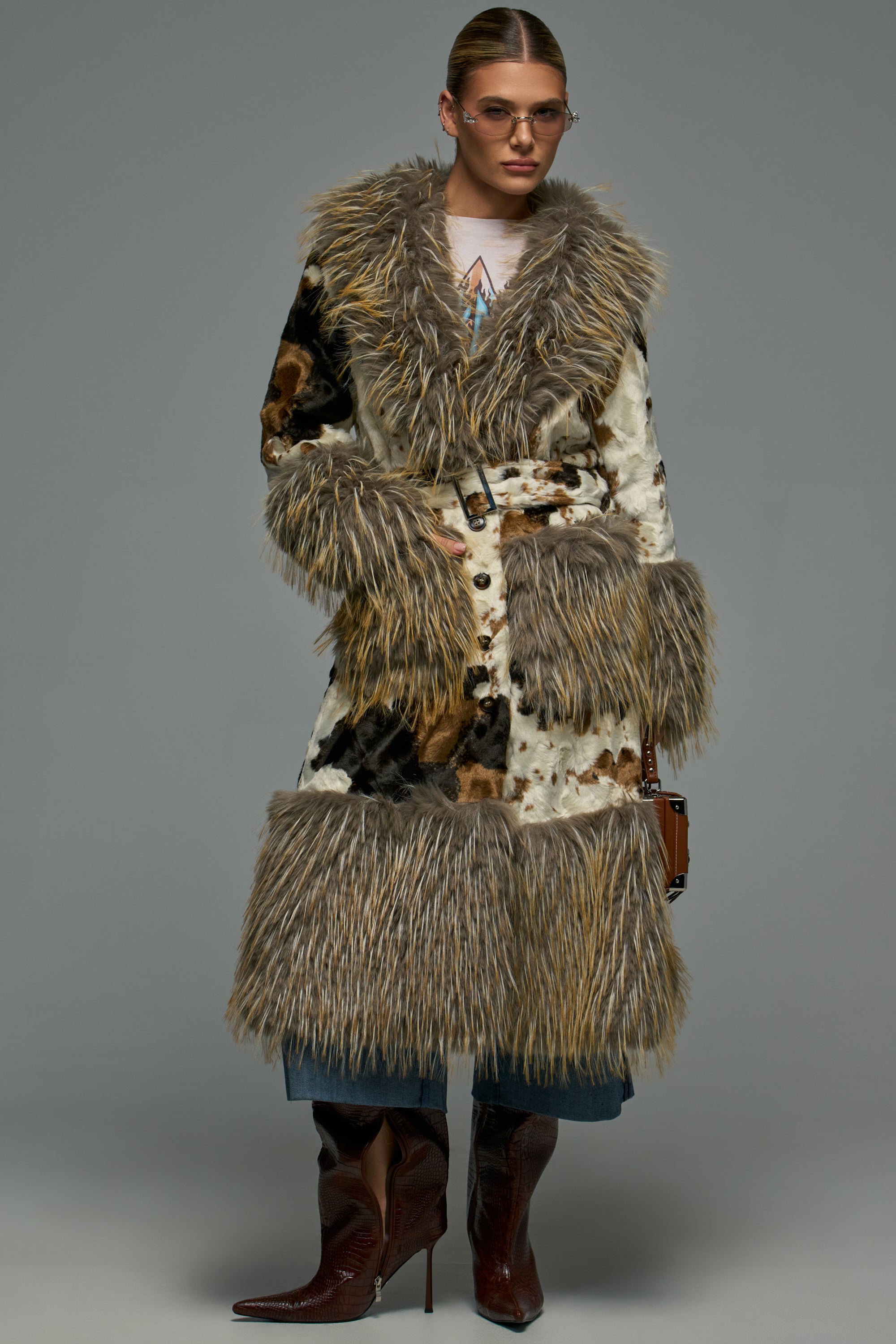 The ROWDY FAUX FUR TRENCH is a belted cow print faux fur coat with shaggy trim on the collar, cuffs, and hem, styled over jeans and dark pointed-toe boots for statement layering.