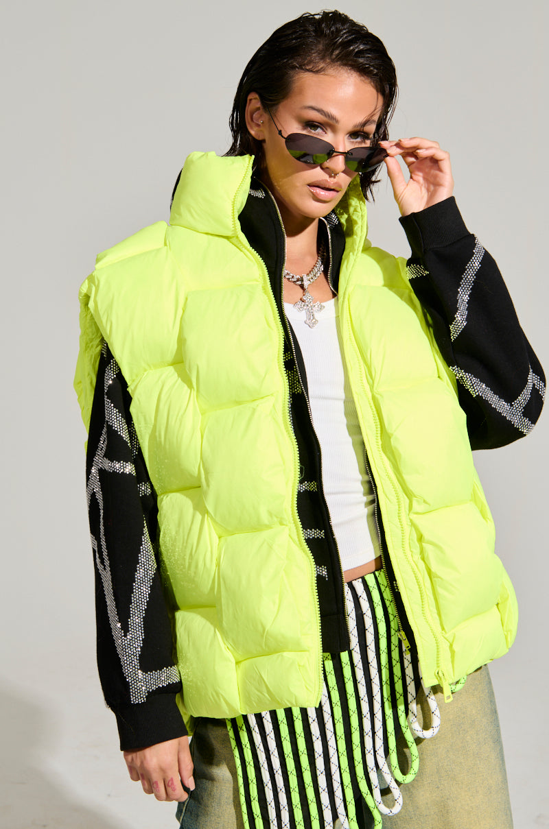 A person poses confidently wearing the LIGHT UP THE NIGHT WEAVE PUFFER VEST in a striking bright yellow, featuring textured fabric and adjustable inner straps, layered over a black and white outfit. They stylishly hold up sunglasses with one hand and sport an edgy hairstyle, all set against a plain background that highlights this bold addition to the outerwear collection.