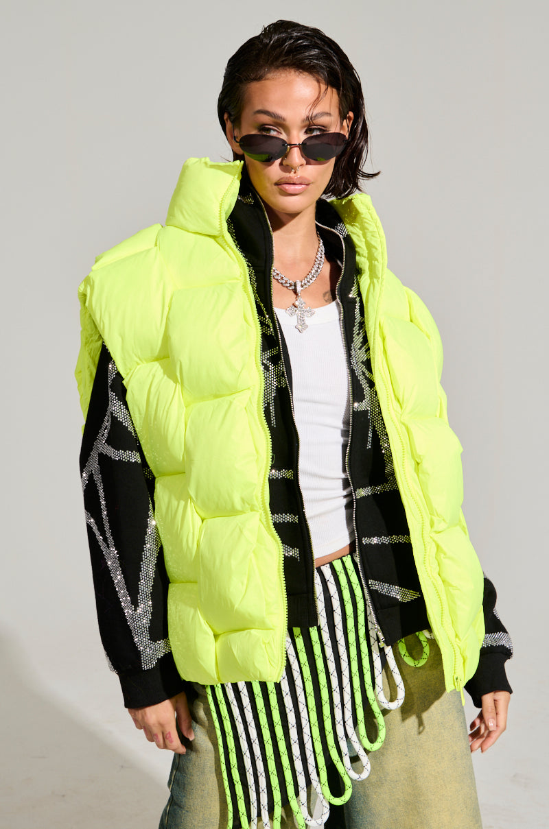A person wearing sunglasses, the "LIGHT UP THE NIGHT WEAVE PUFFER VEST" over a black sweater with silver designs, and green ombre jeans stands confidently. Part of the outerwear collection, this look features short, dark hair for a stylish, modern vibe.