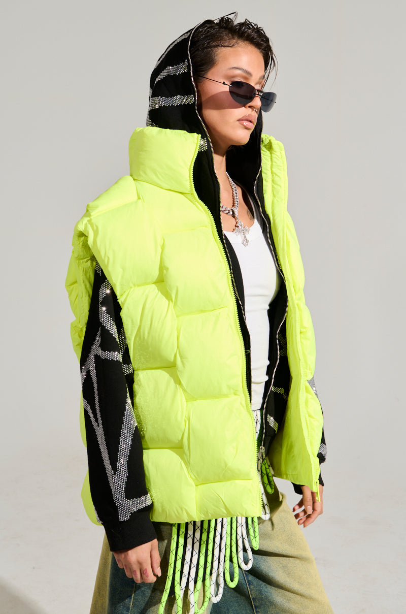 Against a plain backdrop, an individual confidently showcases the LIGHT UP THE NIGHT WEAVE PUFFER VEST in neon yellow over a classic black and white hoodie. With short hair, sunglasses, and jeans featuring fringe details, they embody a trendy urban style ideal for any outerwear collection.