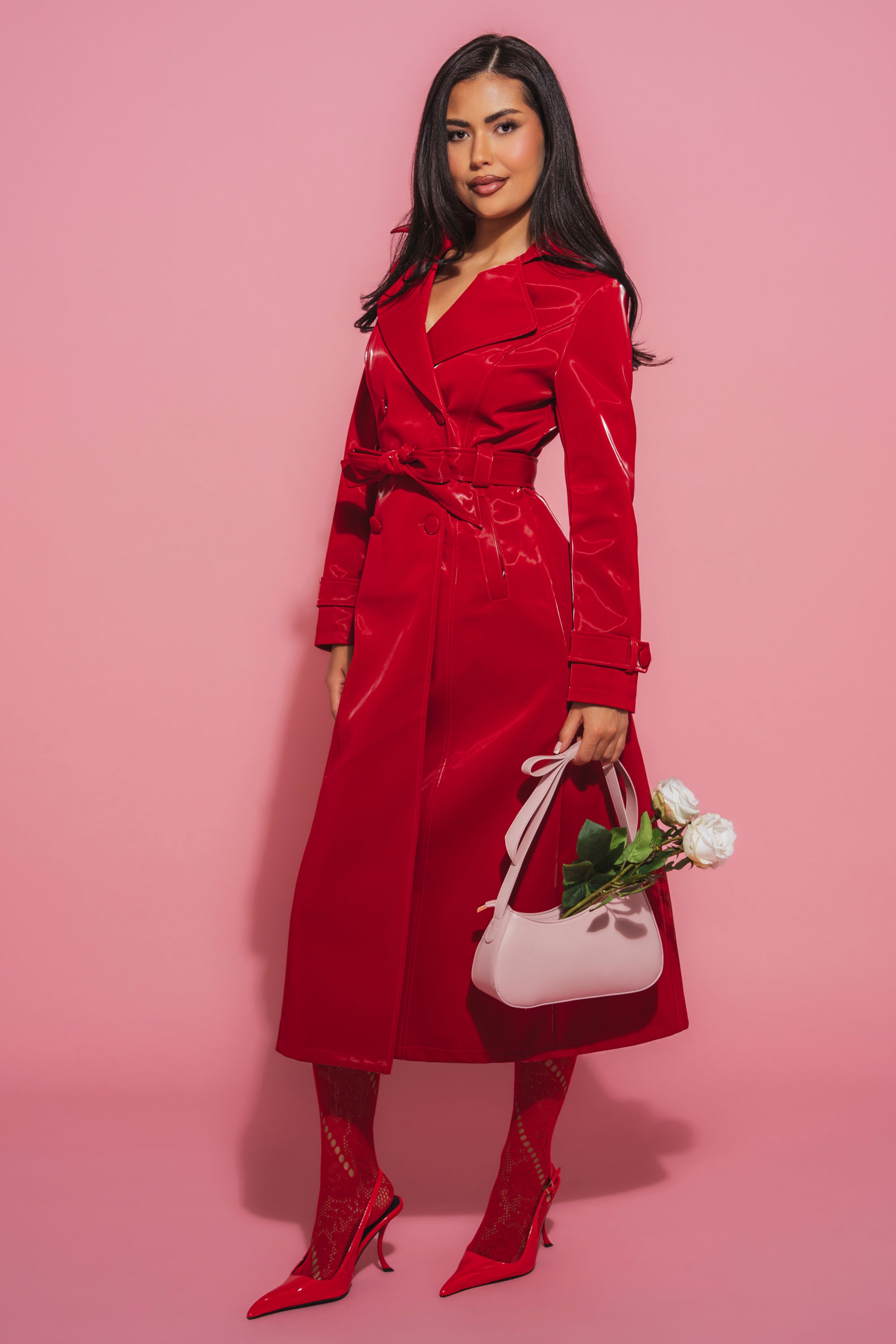 A woman stands against a pink background in the DOZEN ROSES LIQUID VELVET TRENCH, paired with red heels and patterned tights. She holds a pale pink handbag filled with white roses, making a bold style statement. 1
