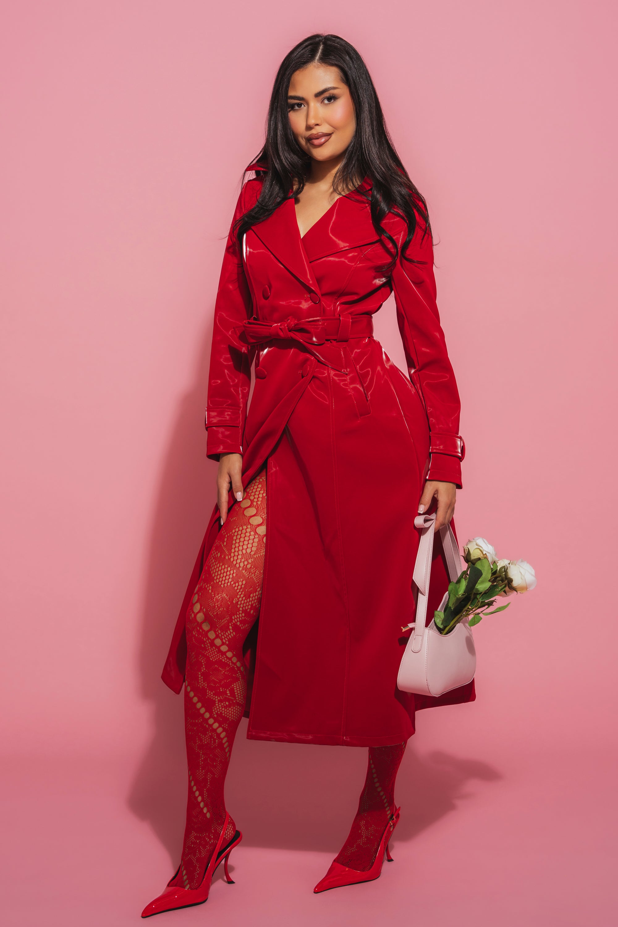 A woman smiles against a pink background in the DOZEN ROSES LIQUID VELVET TRENCH, paired with red patterned tights and heels. She holds a light pink handbag filled with white flowers, her statement outerwear completing the bold look. 2