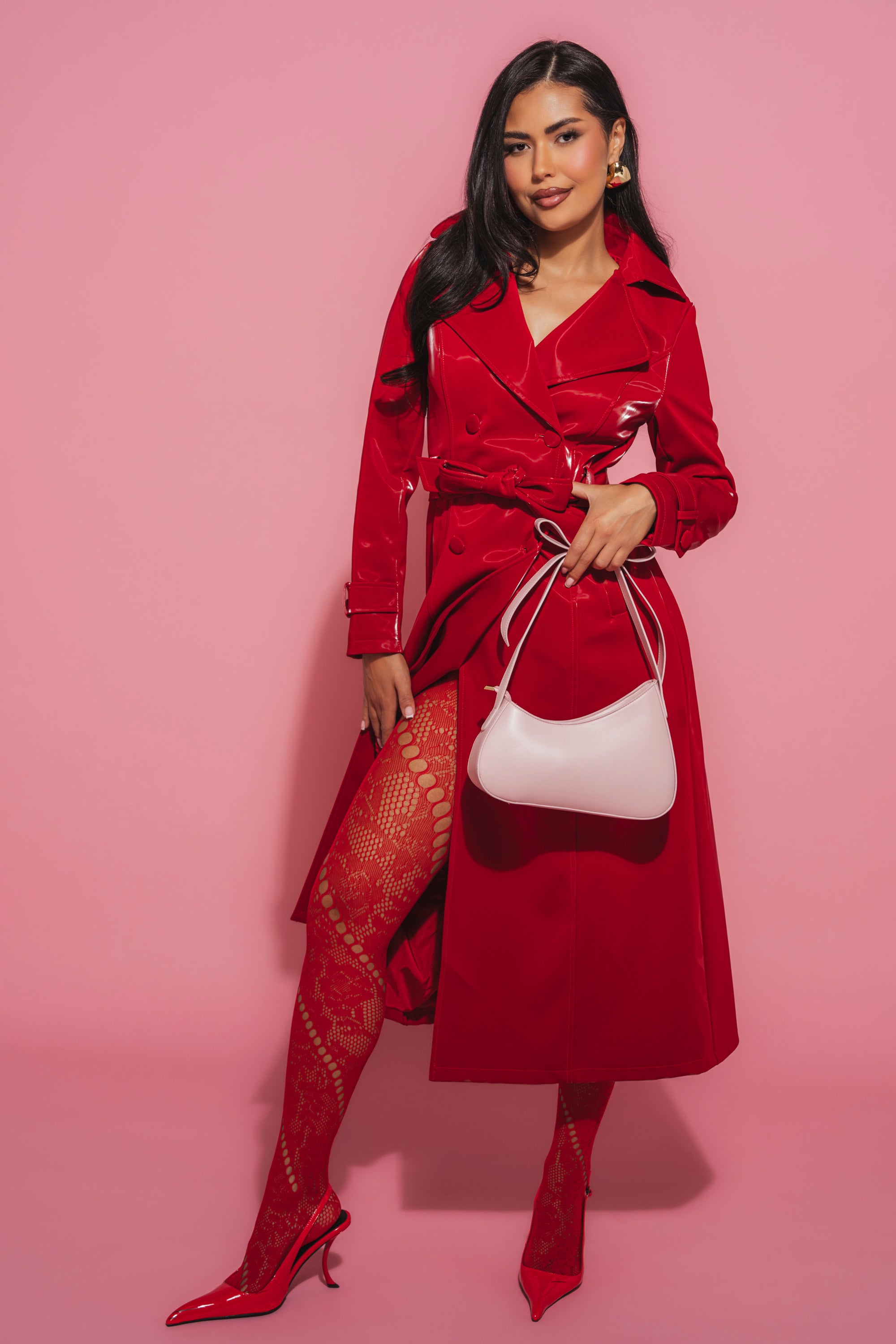 A woman with long dark hair smiles against a pink backdrop, wearing the DOZEN ROSES LIQUID VELVET TRENCH, patterned red tights, red heels, and carrying a white handbag. 3