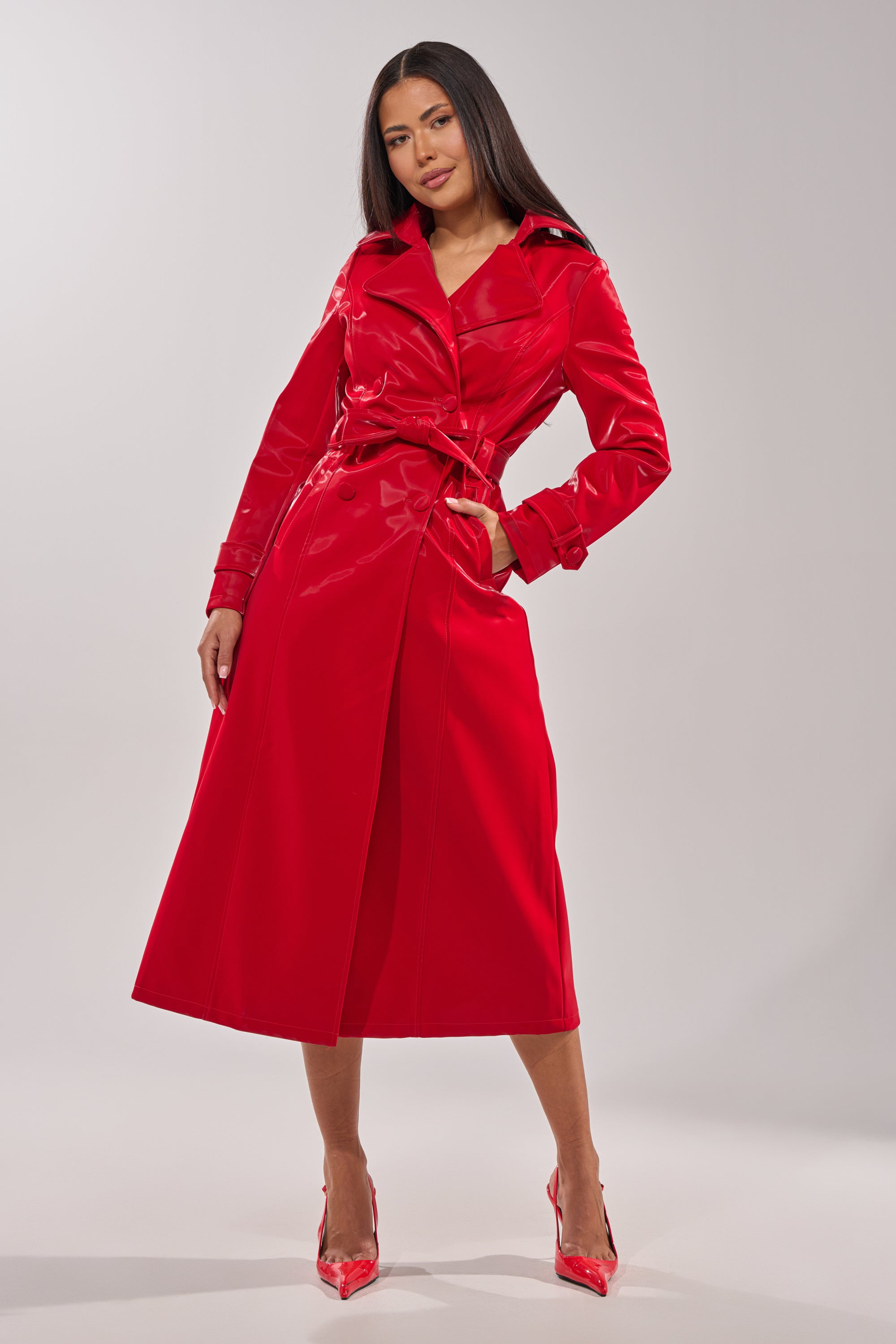 A woman exudes confidence in the DOZEN ROSES LIQUID VELVET TRENCH, a bold red belted coat paired with matching high heels, standing out against a plain light gray background. 8