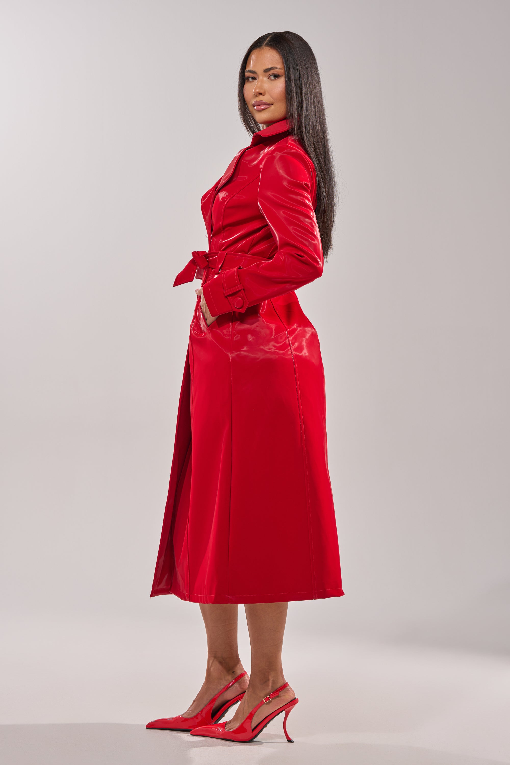A woman with long dark hair stands sideways against a light gray background, wearing the DOZEN ROSES LIQUID VELVET TRENCH and matching red high-heeled shoes. 10