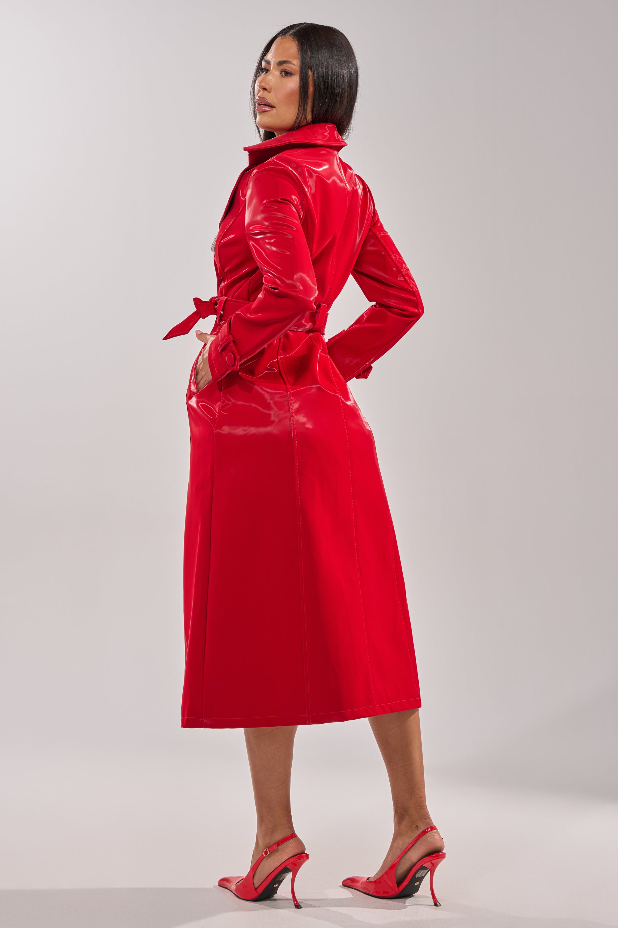A woman models the DOZEN ROSES LIQUID VELVET TRENCH in bright red faux leather, tied at the waist with matching heels. She looks over her shoulder against a light gray backdrop, creating a striking statement outerwear look. 12