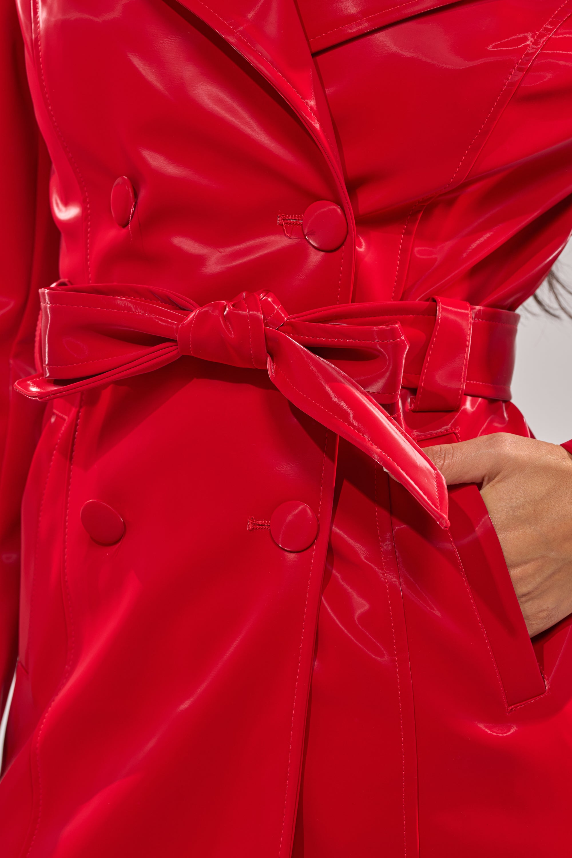 Close-up of the DOZEN ROSES LIQUID VELVET TRENCH, a shiny red faux leather coat with a tie belt and large buttons. A hand in the right pocket highlights this bold outerwear piece. 13