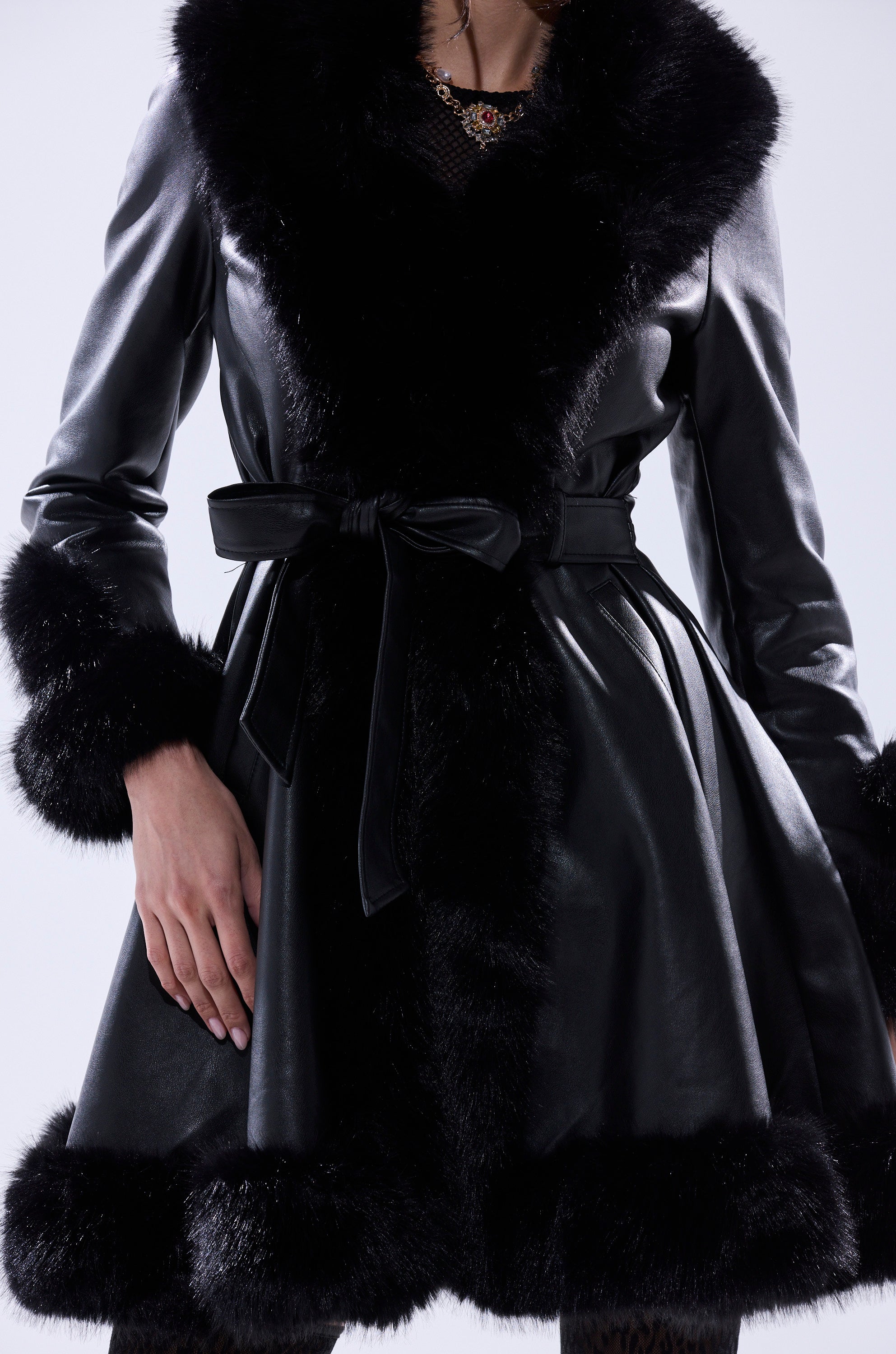 A person wears the ADRIENNE FUR TRIM COAT—a black faux leather jacket with a wide faux fur collar, fur-trimmed cuffs and hem, and a tied waist belt. The image highlights the torso and upper legs, one hand resting on the hip.