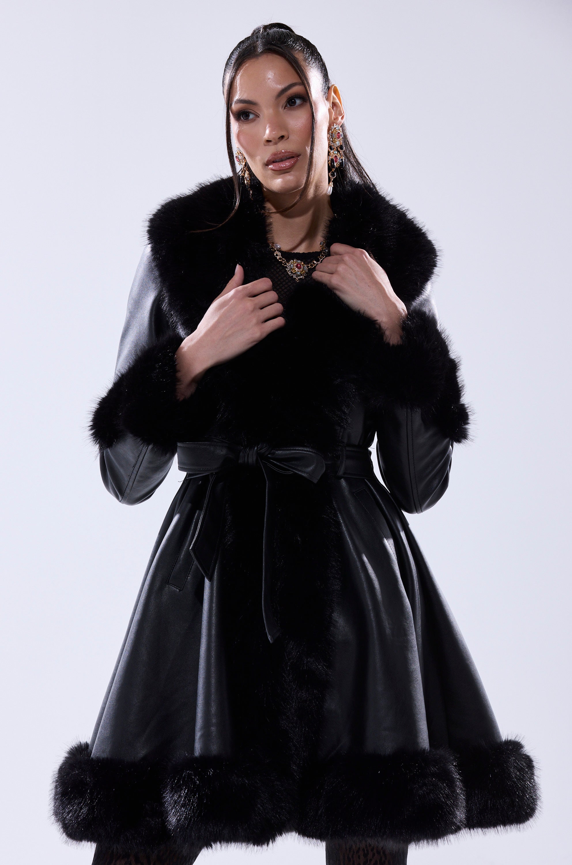 A woman models the ADRIENNE FUR TRIM COAT, featuring a bow belt and plush fur trim, confidently posing against a white background while highlighting the coat’s luxurious texture and elegant style.