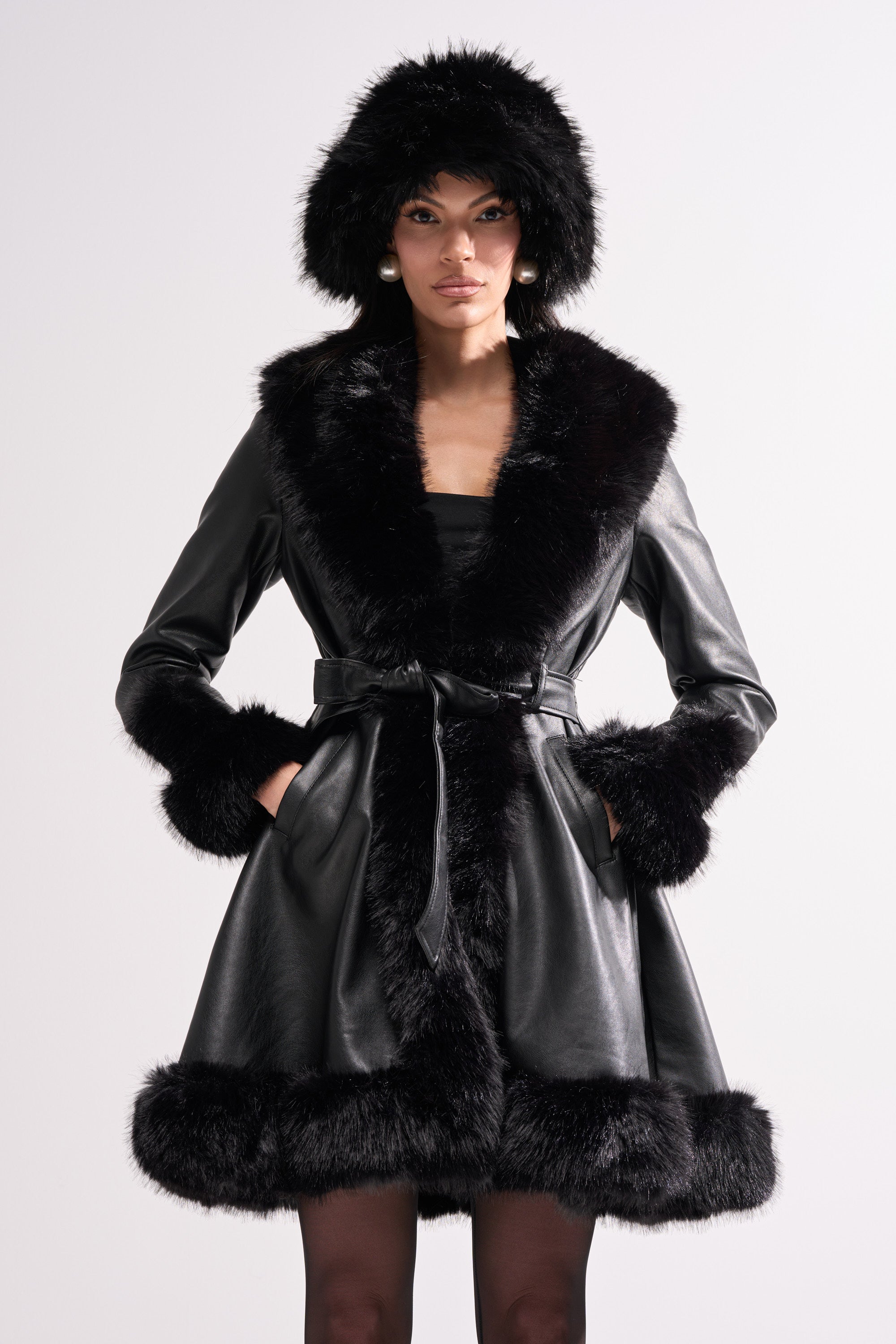A woman models the ADRIENNE FUR TRIM COAT, a chic black faux leather piece with wide fur trim on the collar, cuffs, and hem, plus a belted waist and matching furry hood—ideal statement outerwear for a stylish winter look.