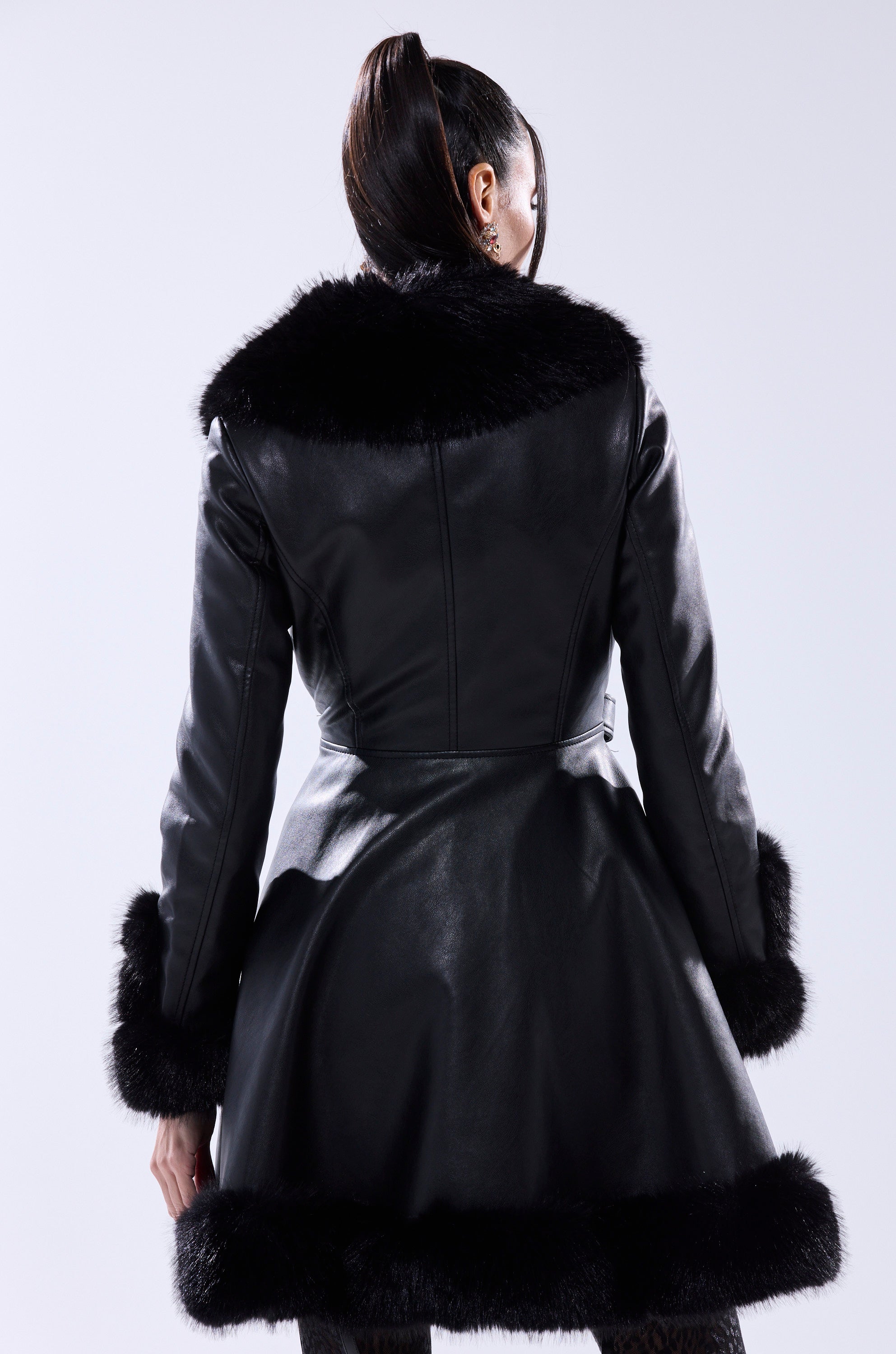 A woman with a high ponytail is seen from behind, wearing the ADRIENNE FUR TRIM COAT—a black faux leather coat with black fur accents on the collar, cuffs, and hem—standing out against a plain light background.