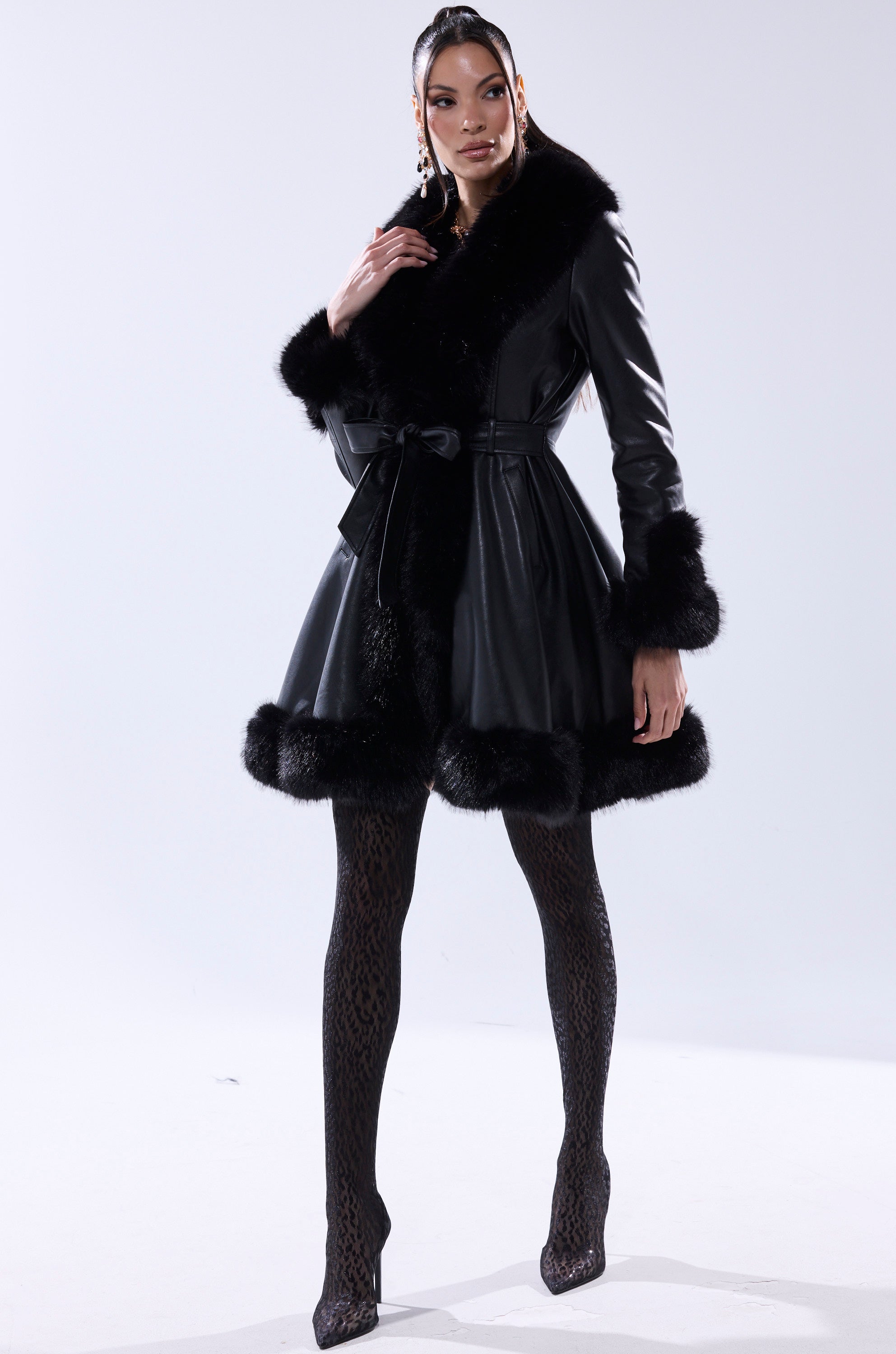 A woman stands confidently in the ADRIENNE FUR TRIM COAT with a waist belt, paired with patterned black tights and high heels against a plain light background.