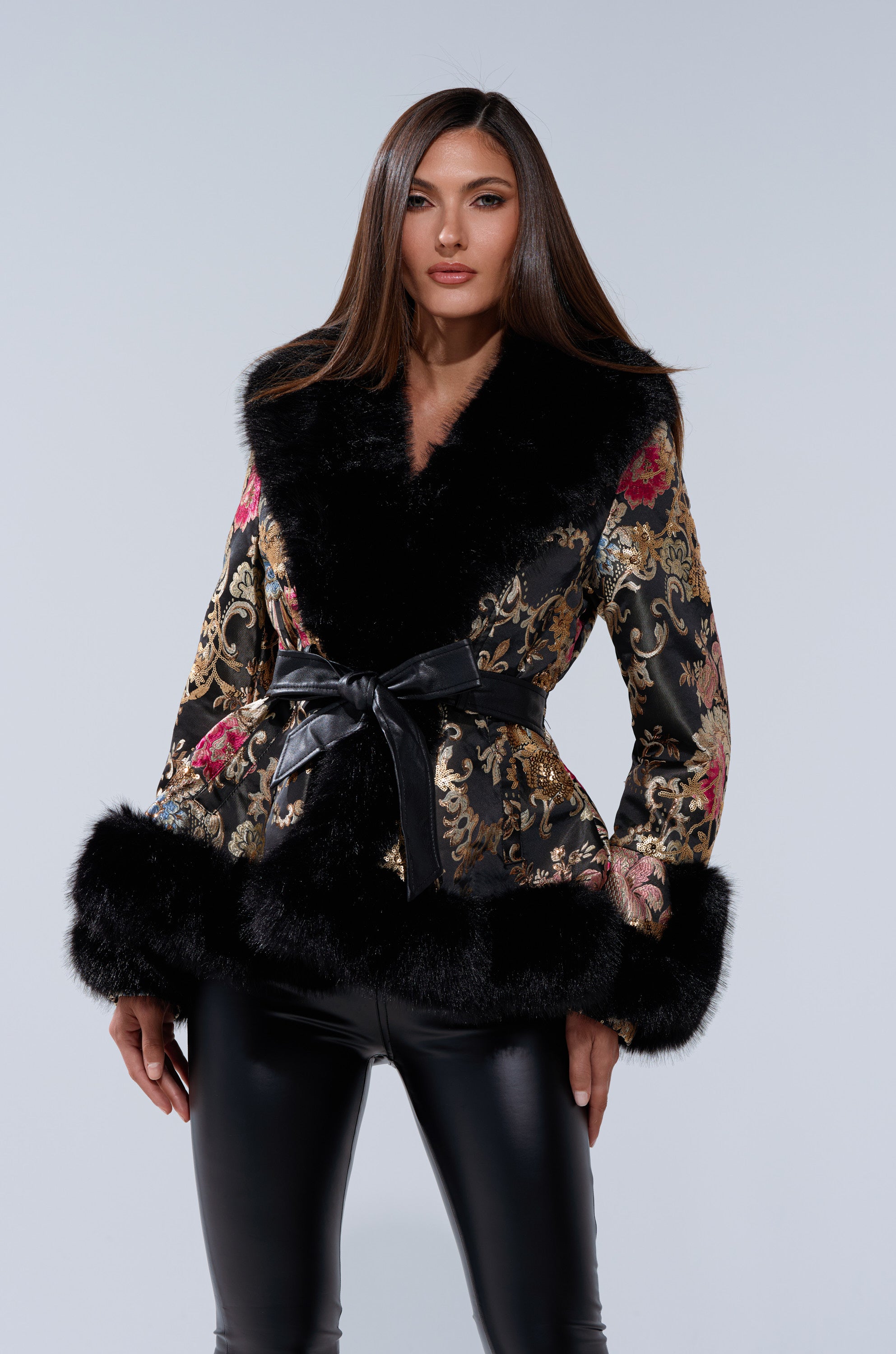 A woman models the KENZY LUXE FUR TRIM MOTO— a black and gold brocade jacket with fur trim and moto details, belted at the waist—paired with black leather pants, set against a plain light gray background.