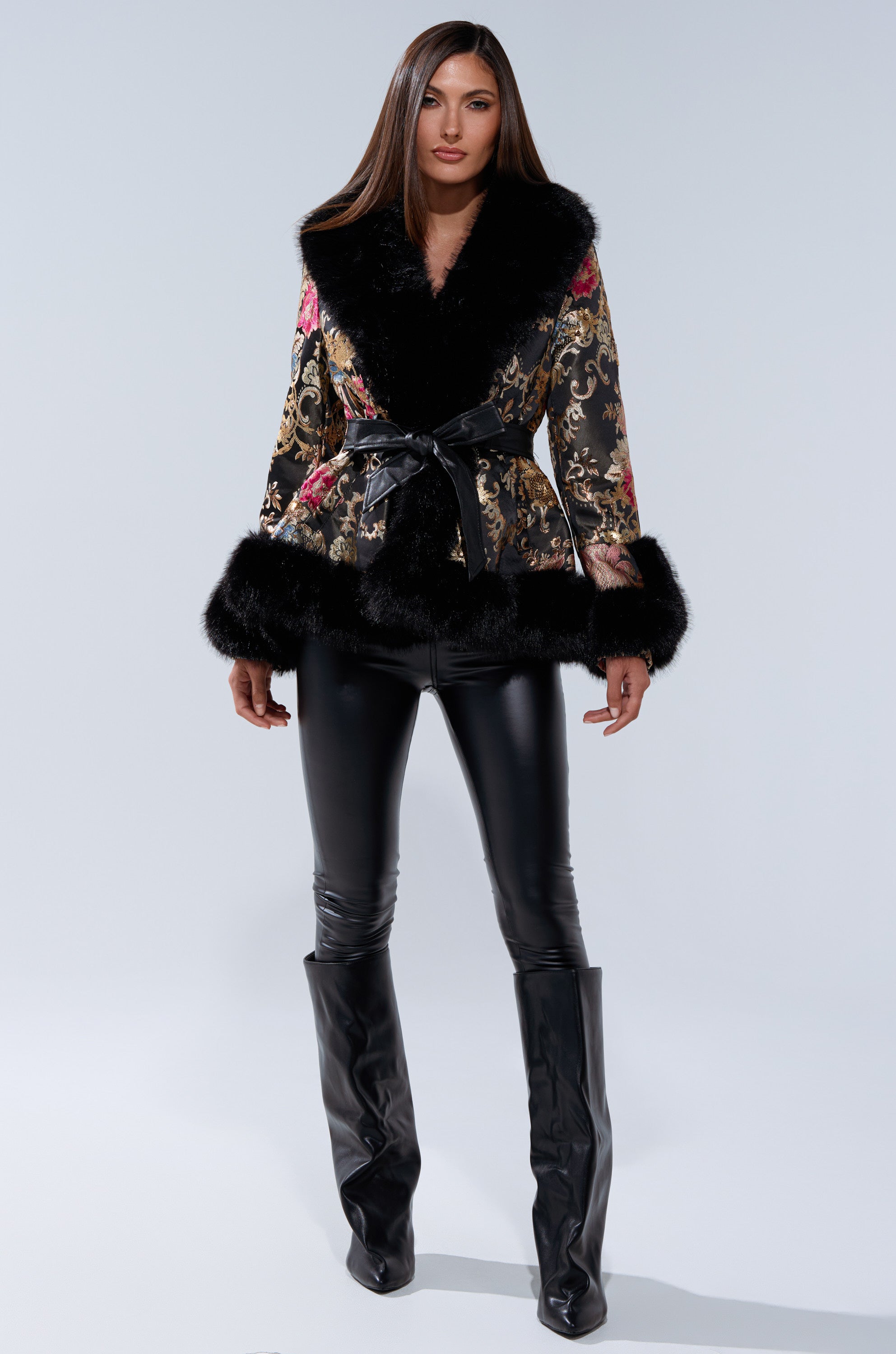 A woman stands confidently in the KENZY LUXE FUR TRIM MOTO, a black brocade coat with fur trim, styled with black leather pants and knee-high boots against a simple light backdrop.