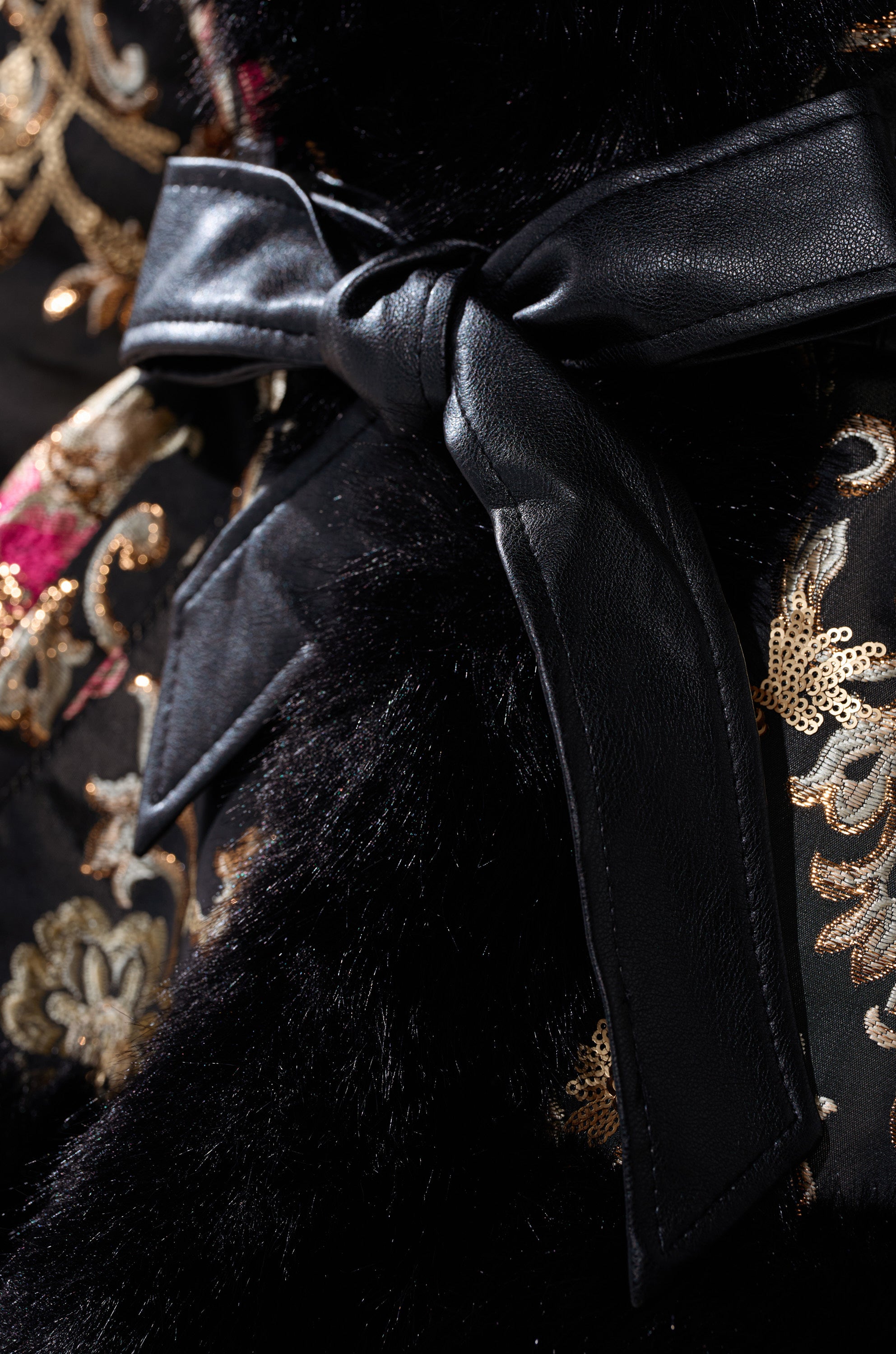 Close-up of the KENZY LUXE FUR TRIM MOTO jacket with a black leather belt tied in a bow, detailed black fur trim, and intricate gold and red floral brocade embroidery.