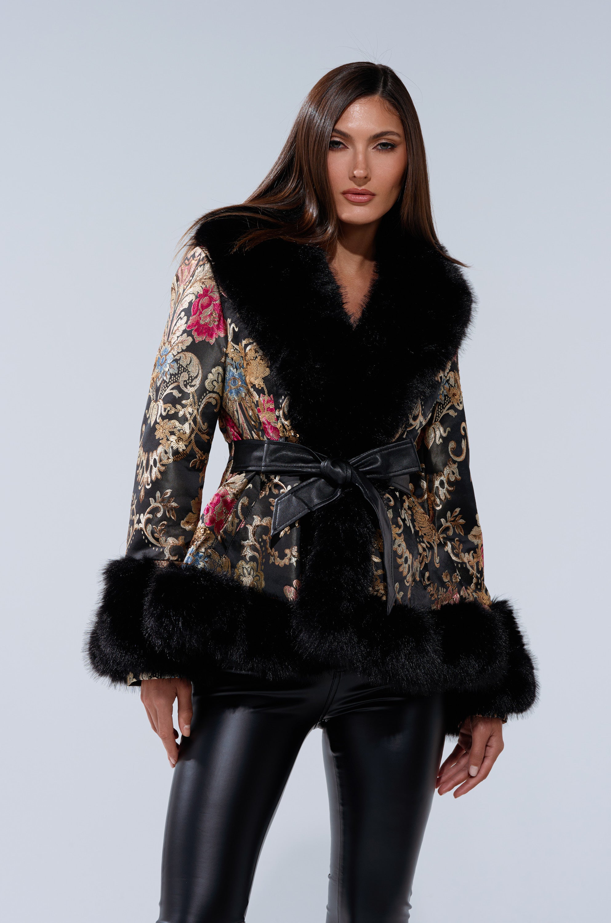 A woman wears the KENZY LUXE FUR TRIM MOTO black brocade jacket with fur trim and a belt, paired with black leather pants, standing against a plain light gray background.