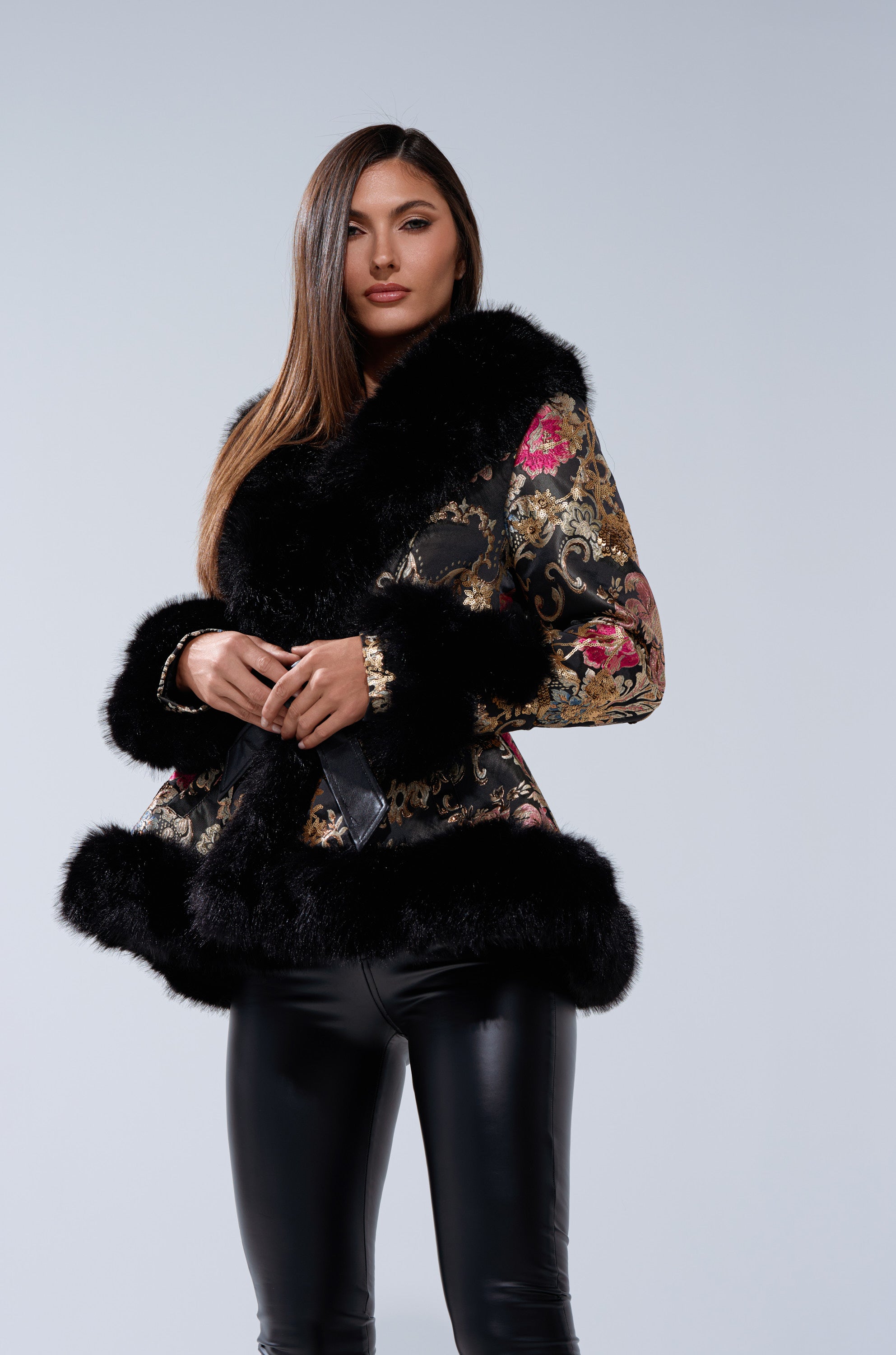A woman stands confidently against a light gray background, wearing the KENZY LUXE FUR TRIM MOTO—a black and gold brocade coat with black fur trim—paired with black leather pants.