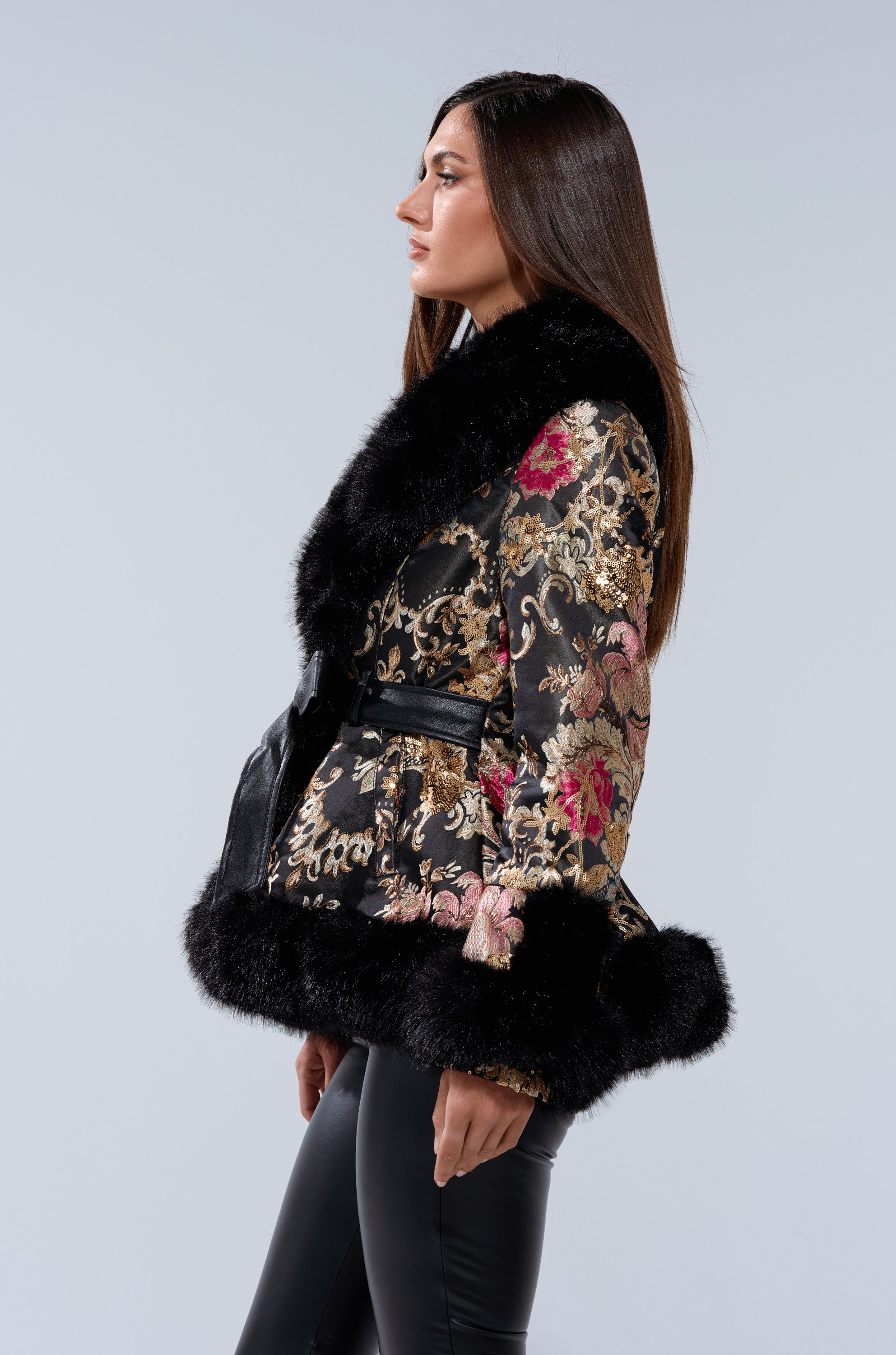 A woman in profile wears the KENZY LUXE FUR TRIM MOTO, a black and gold brocade coat with pink accents and black faux fur trim at the collar and hem, paired with black leather pants against a neutral backdrop.