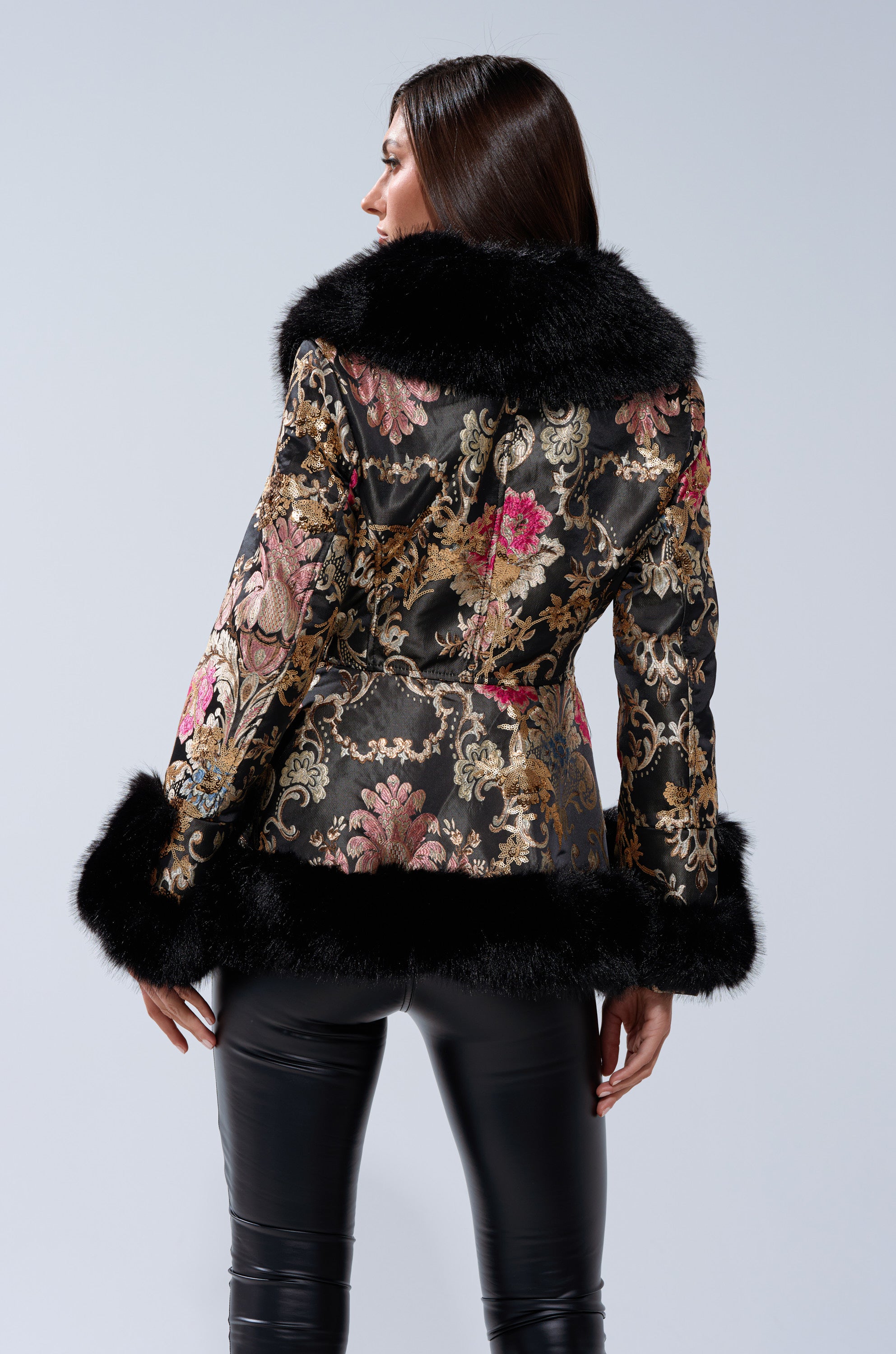 A woman faces away, wearing the KENZY LUXE FUR TRIM MOTO— a black brocade coat with fur-trimmed collar, cuffs, and hem—paired with black leather pants against a neutral background.