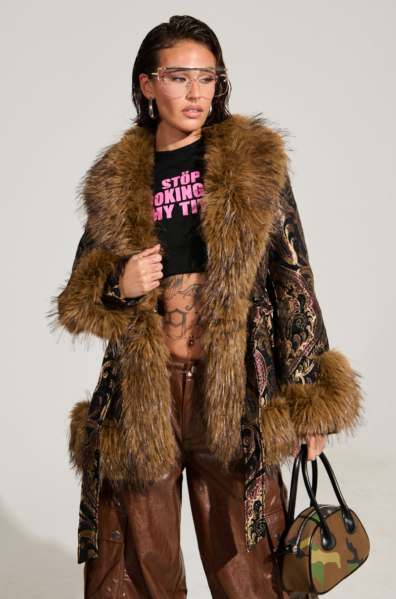 Wearing the chic SITTING PRETTY FUR TRIM COAT, a person poses confidently in stylish sunglasses. The coat, adorned with faux fur cuffs, is layered over a black crop top paired with brown leather pants. They carry a small camouflage bag, with the bold pink text on the top beautifully complementing the overall look.