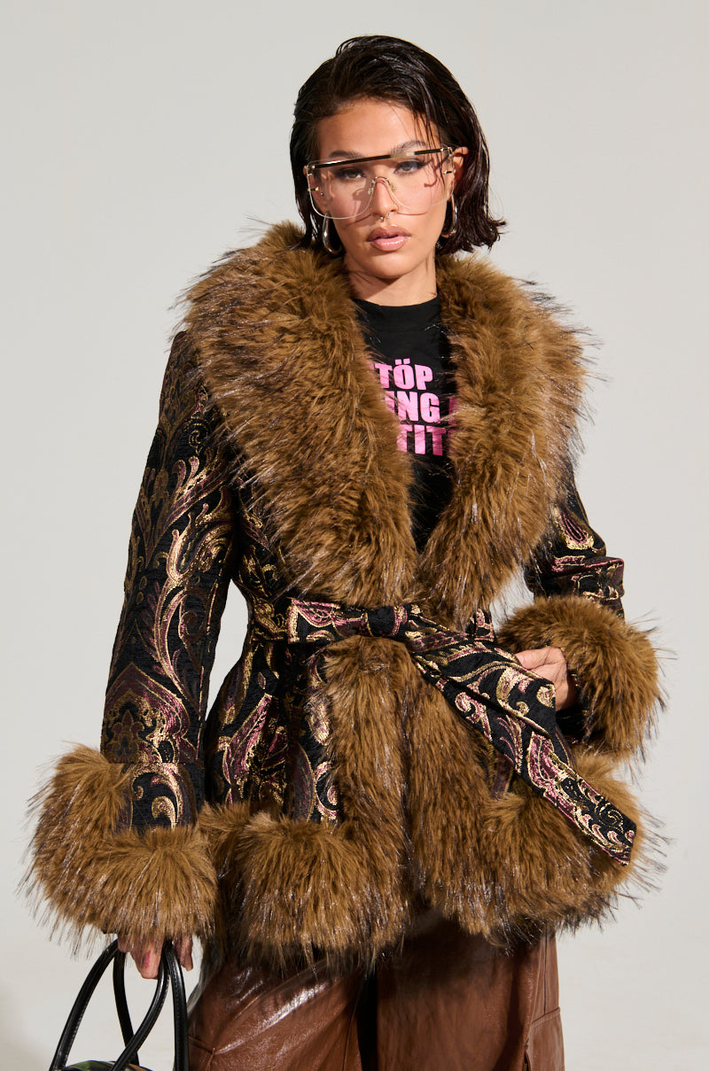 Wearing the SITTING PRETTY FUR TRIM COAT, featuring a lavish design with velvet fabric and faux fur cuffs, a person pairs it with brown pants and aviator-style glasses while holding a black bag. The coat is accentuated by its belt and large fur collar. With a neutral expression, the person gazes ahead.