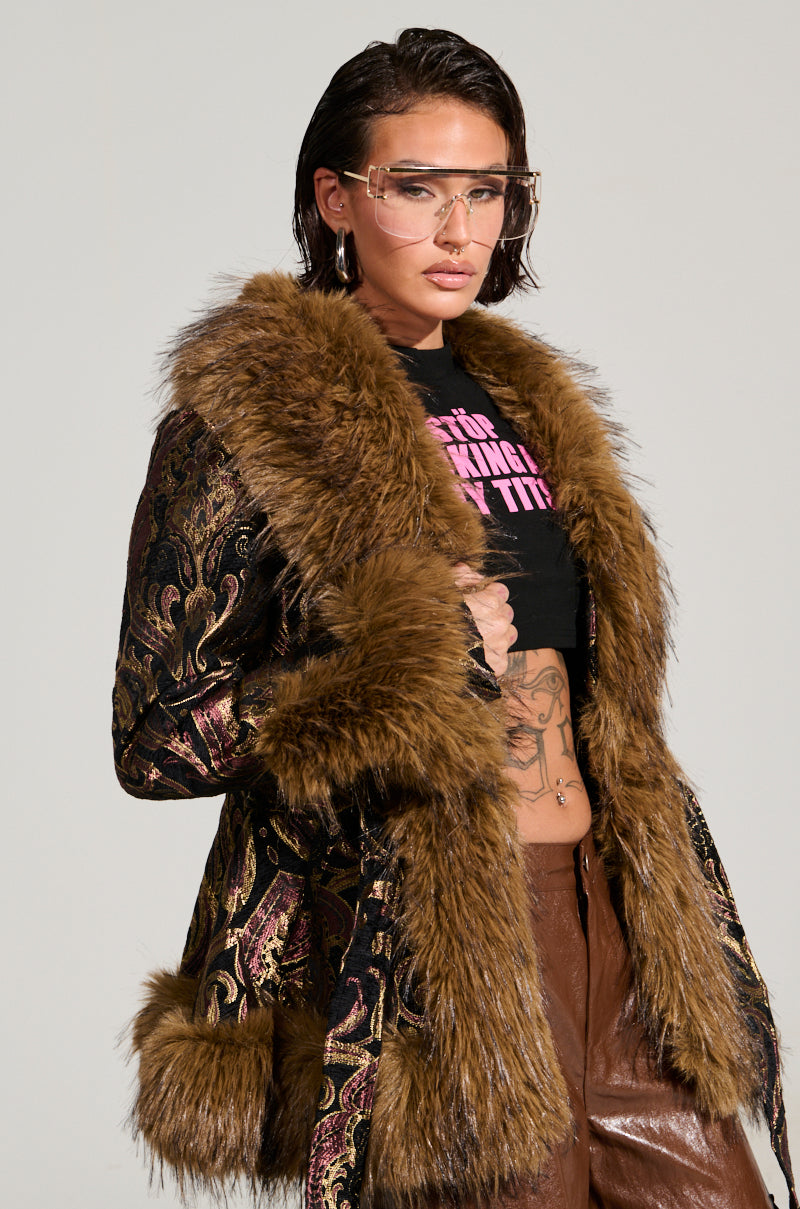Wearing the SITTING PRETTY FUR TRIM COAT with faux fur cuffs, a person is styled in brown pants and a graphic black crop top. Their short hair and clear visor-style glasses complement the look, with tattoos visible on their torso.