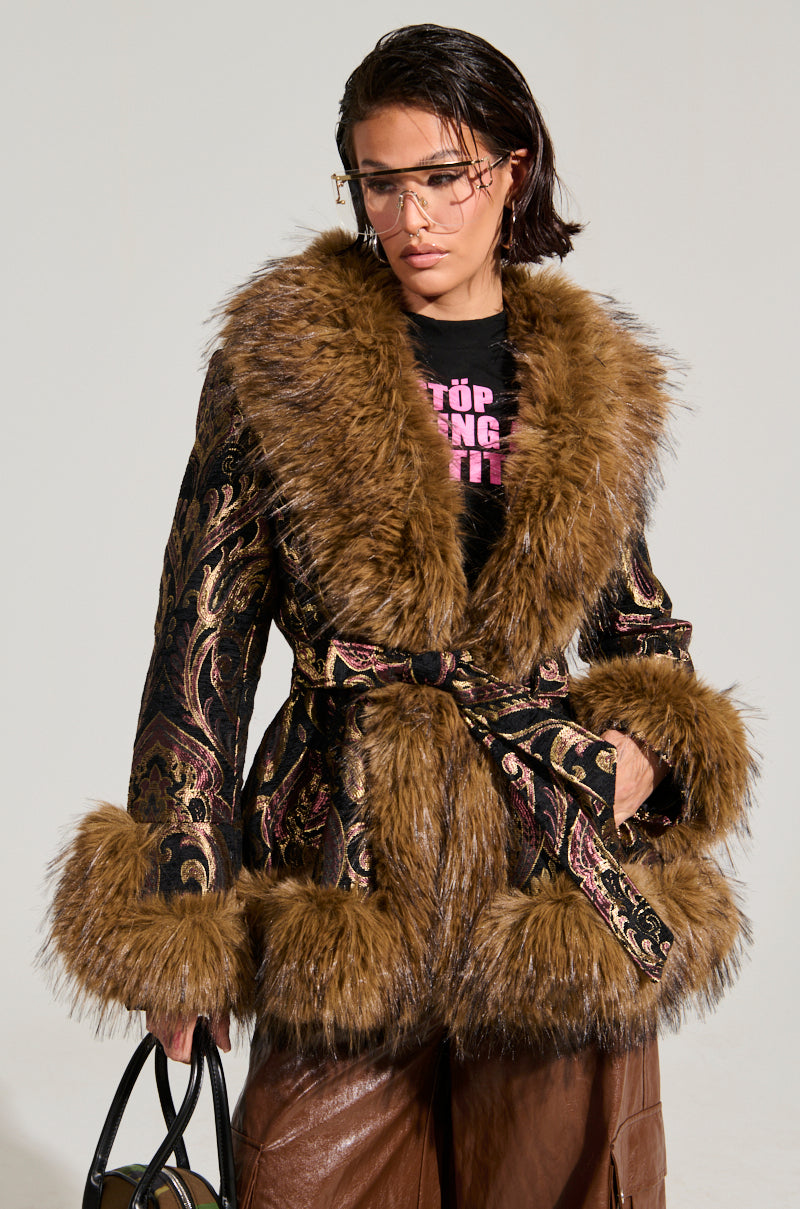 A person wearing the elegant SITTING PRETTY FUR TRIM COAT, featuring a black and gold patterned design with large brown faux fur cuffs, stands against a light background. They have short dark hair and wear clear eyeglasses. The belted coat enhances their elegance, and they hold a small black handbag to complete the chic ensemble.