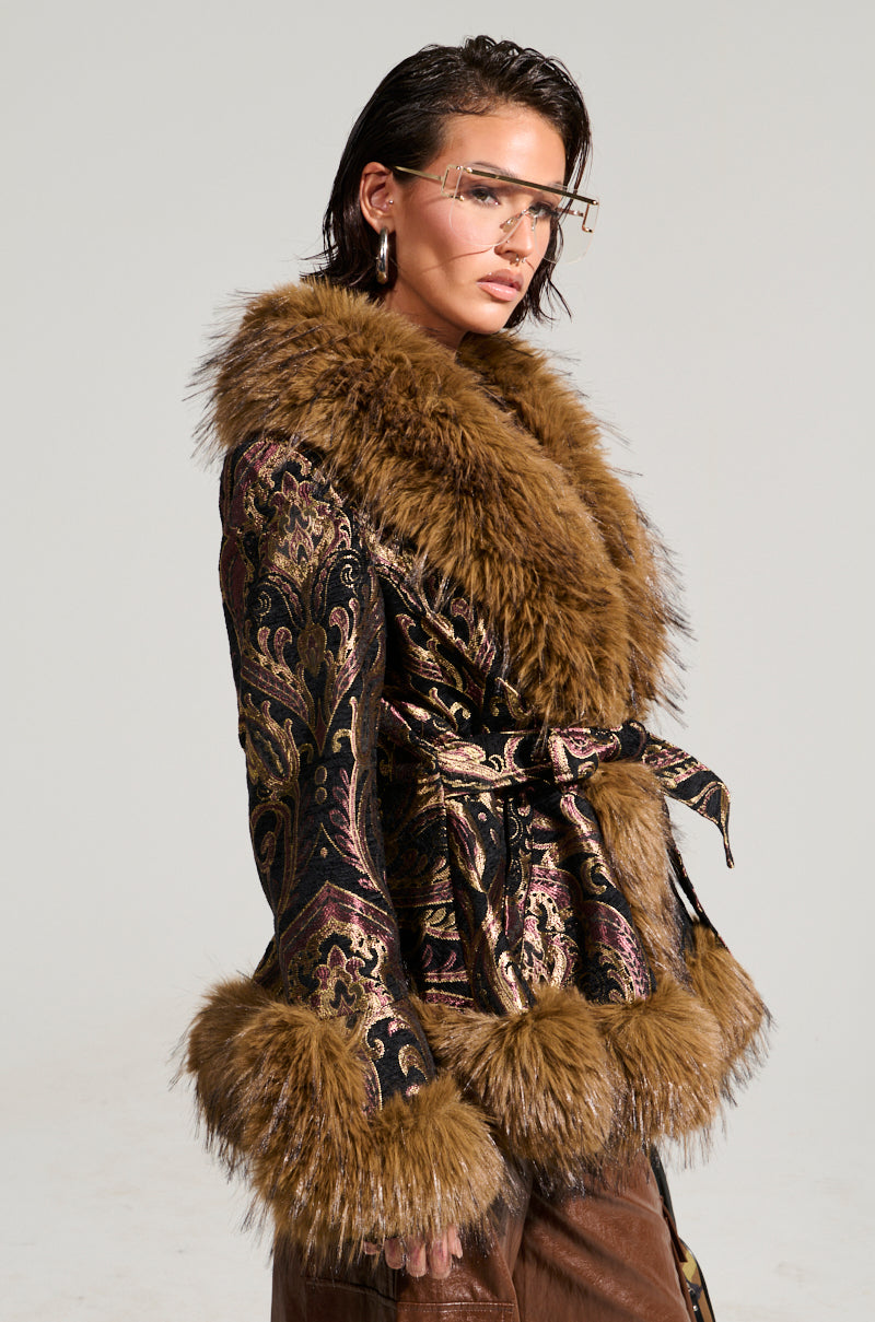 Dressed confidently in the stylish SITTING PRETTY FUR TRIM COAT, an individual showcases the luxurious velvet fabric with faux fur trim. The design's rich brown and gold tones perfectly complement the faux fur collar and cuffs. Their face is adorned with large, fashionable glasses, and their slicked-back hair stands out against a simple backdrop.