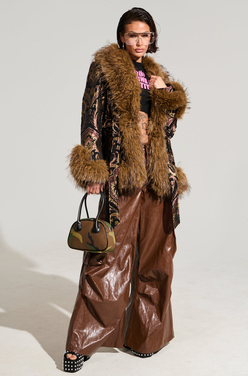 Someone poses confidently in the SITTING PRETTY FUR TRIM COAT, paired with leather pants and sunglasses. The coat's faux fur cuffs add a touch of elegance. They hold a small camouflage-patterned handbag against the simple, light-colored background.