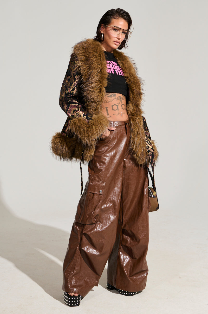A person poses confidently in a black top and the "Sitting Pretty Fur Trim Coat," adorned with intricate designs. Their ensemble includes brown wide-leg leather pants, large glasses, and studded platform shoes, highlighted by velvet fabrications that underscore their bold fashion statement.