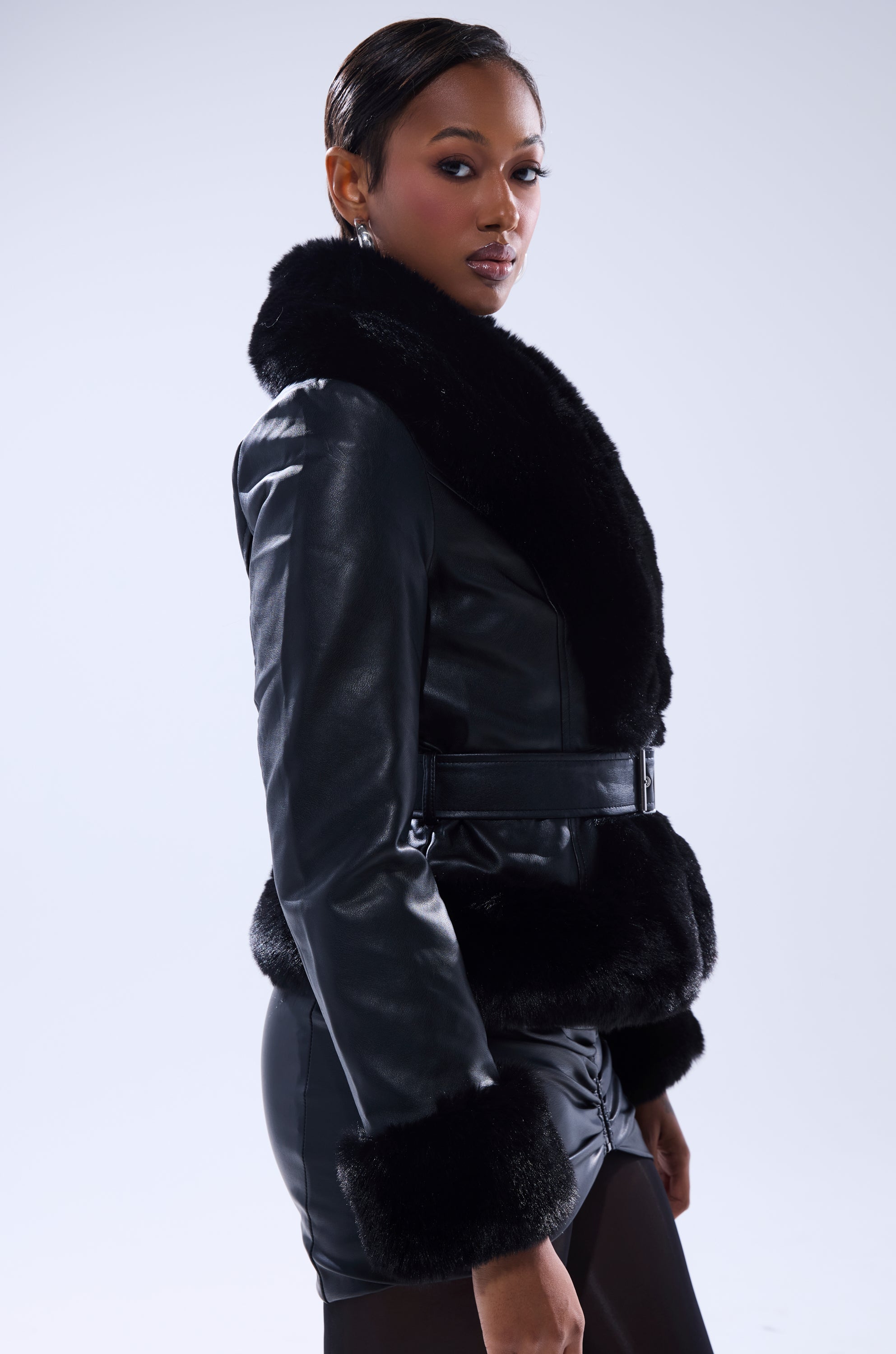 A woman models the KYLA FUR TRIM MOTO faux leather jacket with a longline silhouette, confidently posing against a light gray background with her sleek hair pulled back as she gazes over her shoulder. 2