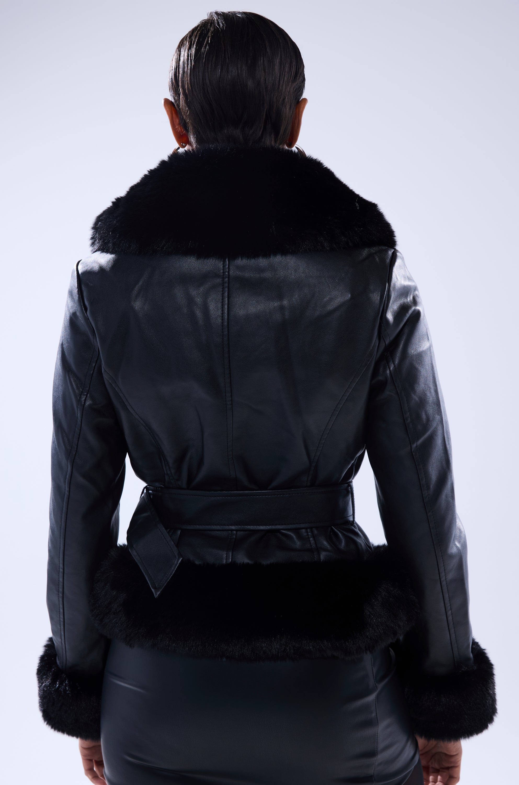 Seen from behind, a person with short, slicked-back hair wears the KYLA FUR TRIM MOTO—a black faux leather jacket featuring a wide, fluffy fur collar and cuffs plus a belted waist—against a plain light gray background. 3