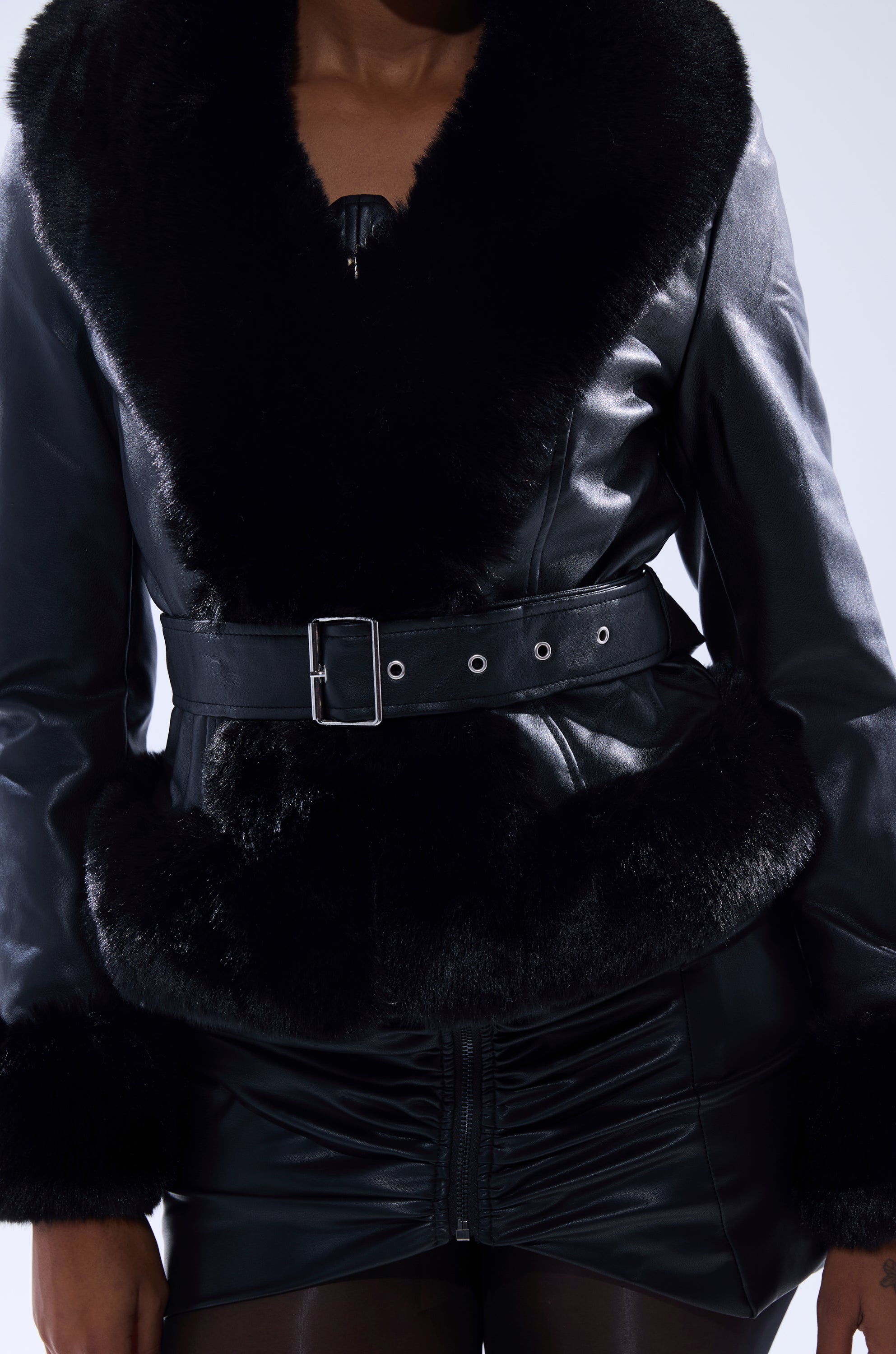 A person wears the KYLA FUR TRIM MOTO, a black faux leather moto jacket with fur trim and belted waist, paired with a ruched black leather skirt. Only the torso and upper legs are shown. 6