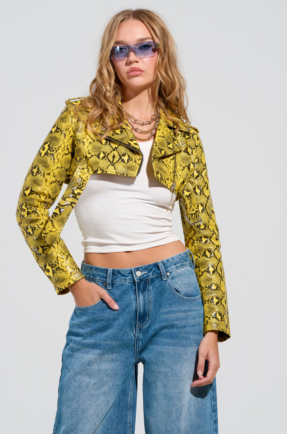 A person with long hair wears a yellow faux snakeskin moto jacket called the SYLVETTE ULTRA CROP MOTO, along with a white crop top, blue jeans, and blue sunglasses. They stand with one hand in their pocket against a plain background.