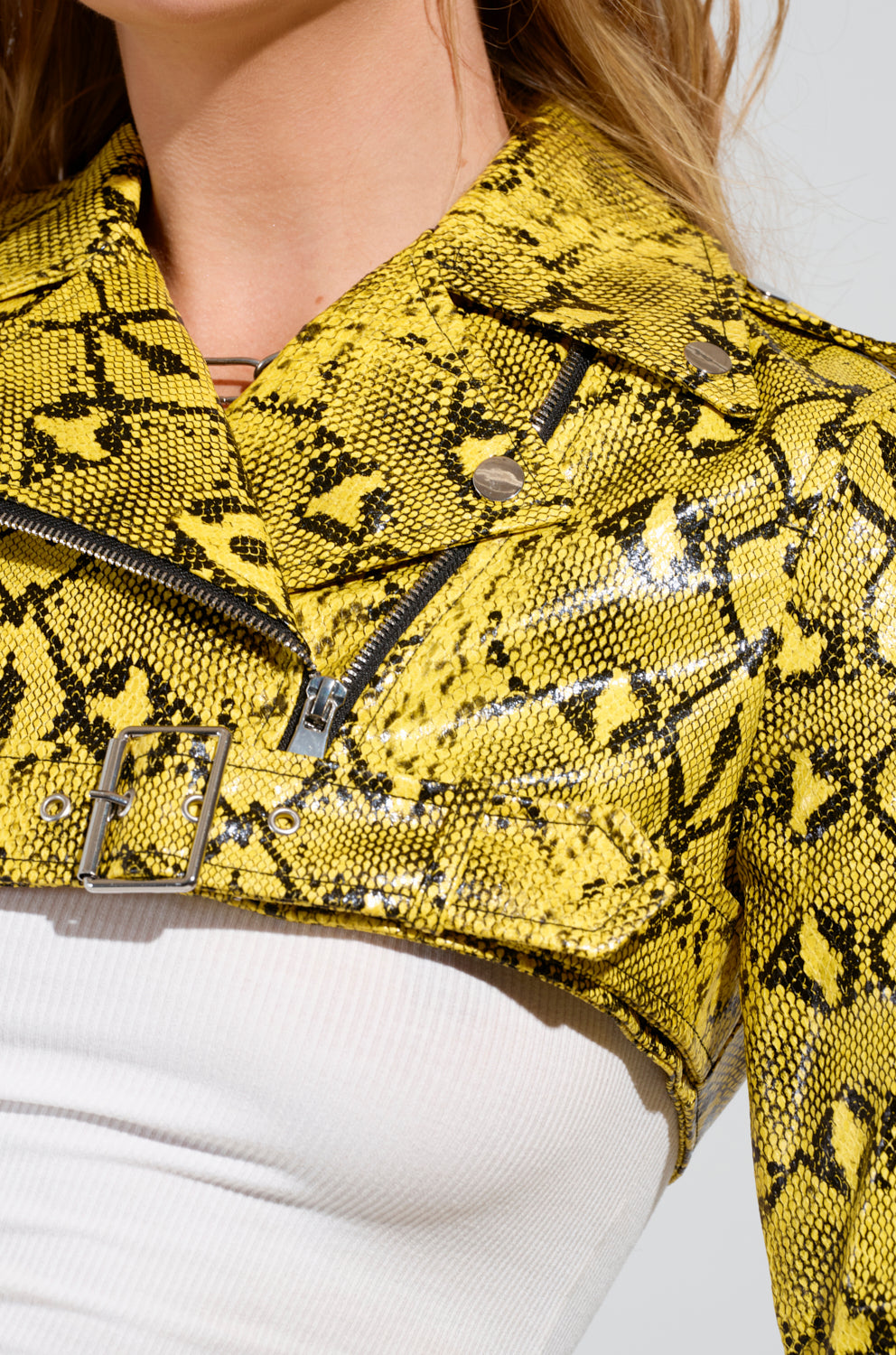 Wearing the eye-catching SYLVETTE ULTRA CROP MOTO jacket in faux snakeskin, featuring zippers, silver studs, and a belt detail, a person layers it over a white ribbed tank top.