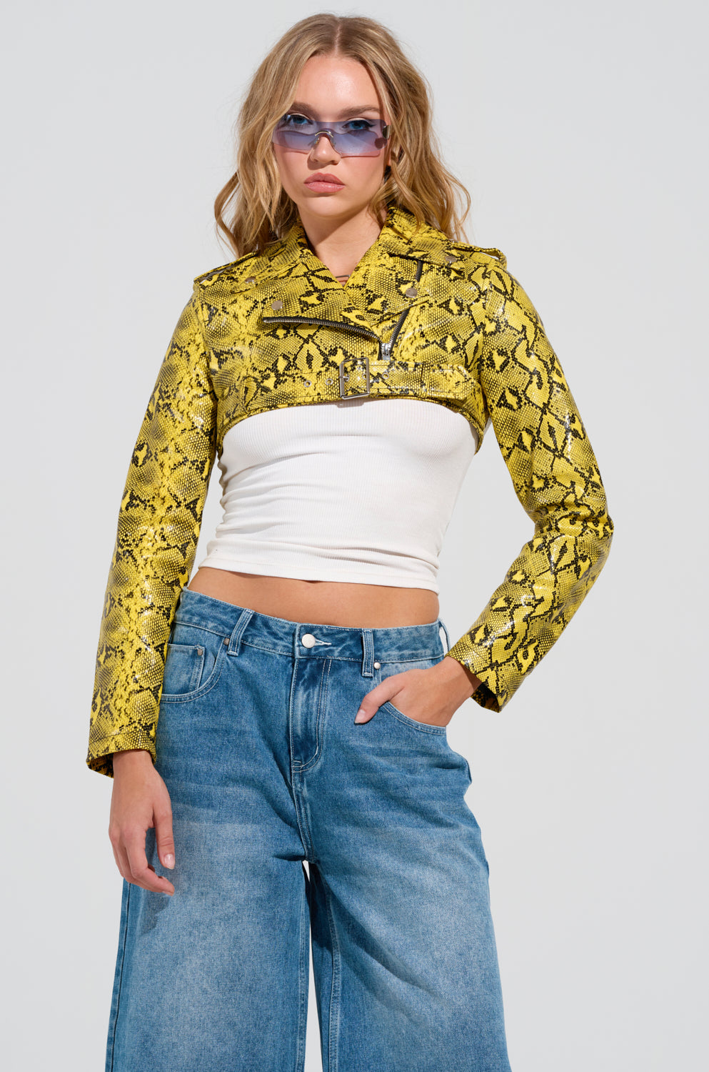 A person sporting the SYLVETTE ULTRA CROP MOTO jacket in faux snakeskin effortlessly poses against a plain background. They wear it over a white crop top paired with wide-leg jeans, accessorized with sunglasses and long wavy hair, while one hand is casually tucked in their pocket.