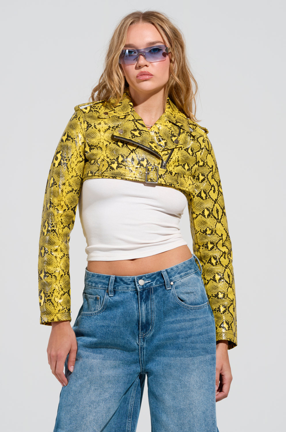 Sporting the SYLVETTE ULTRA CROP MOTO jacket, which boasts a striking faux snakeskin pattern in yellow, a person stands confidently. They complement the look with a white sleeveless top, blue jeans, and blue-tinted sunglasses while their long hair elegantly falls against the plain white background.